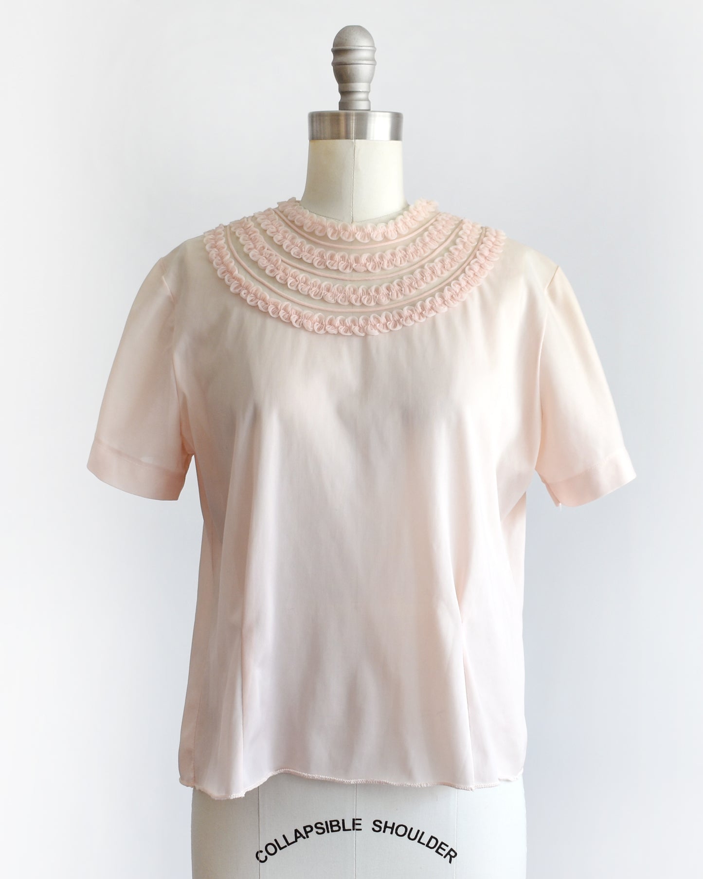 A vintage 1960s pale blush pink short-sleeve blouse with a decorative layered ruffle collar, displayed on a white mannequin dress form against a white background.