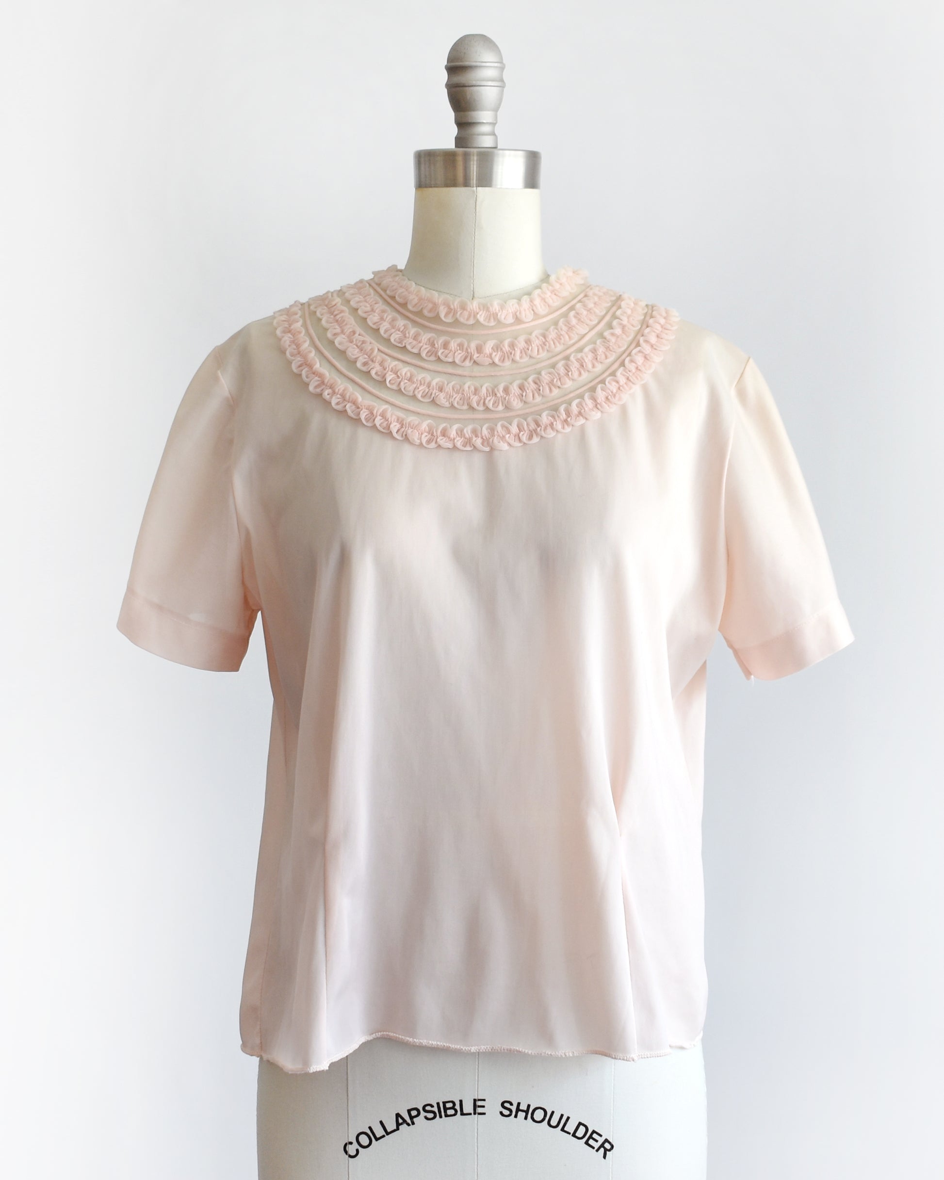 A vintage 1960s pale blush pink short-sleeve blouse with a decorative layered ruffle collar, displayed on a white mannequin dress form against a white background.