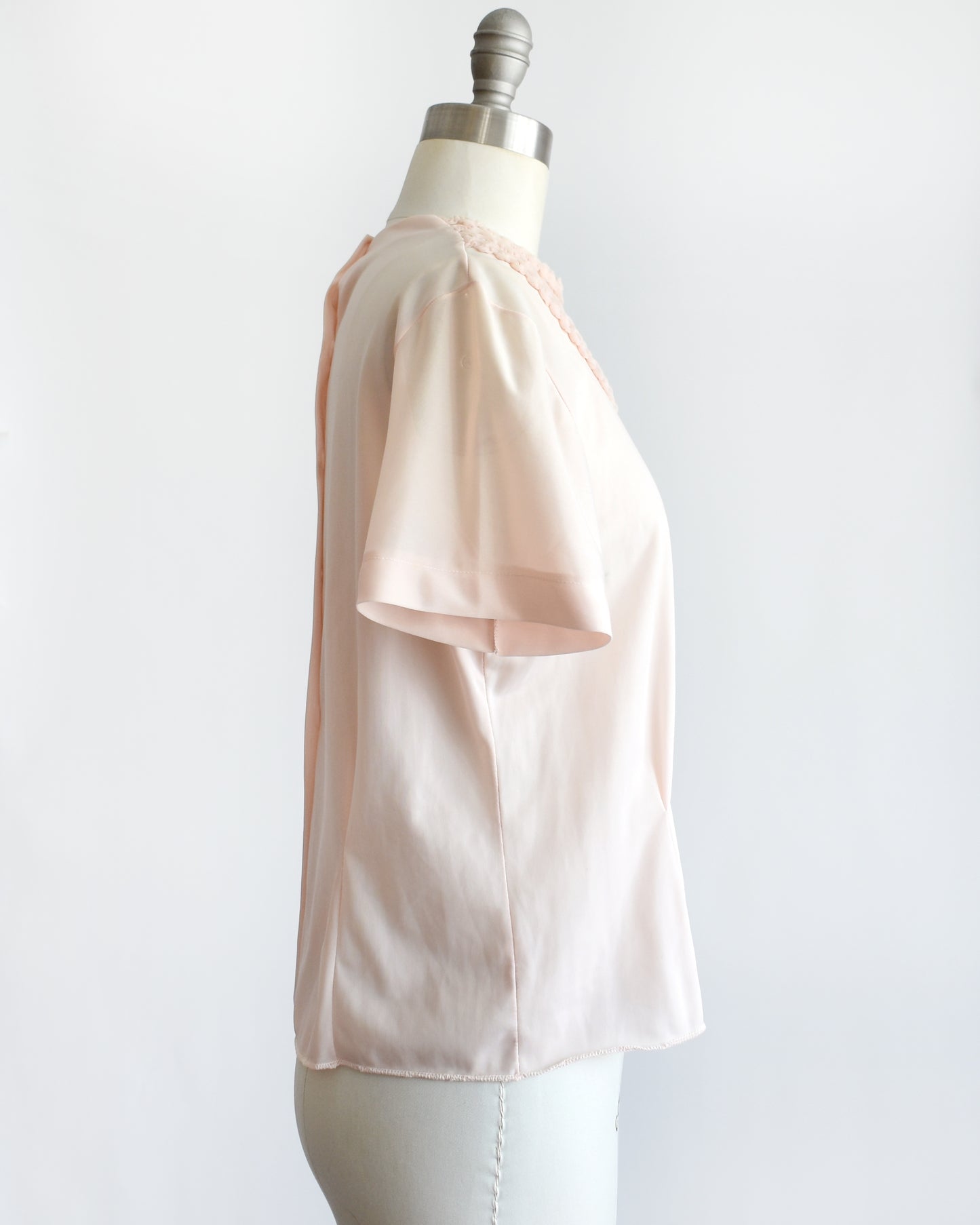 side view of a vintage 1960s pale blush pink short-sleeve blouse with a decorative layered ruffle collar, displayed on a white mannequin dress form against a white background.