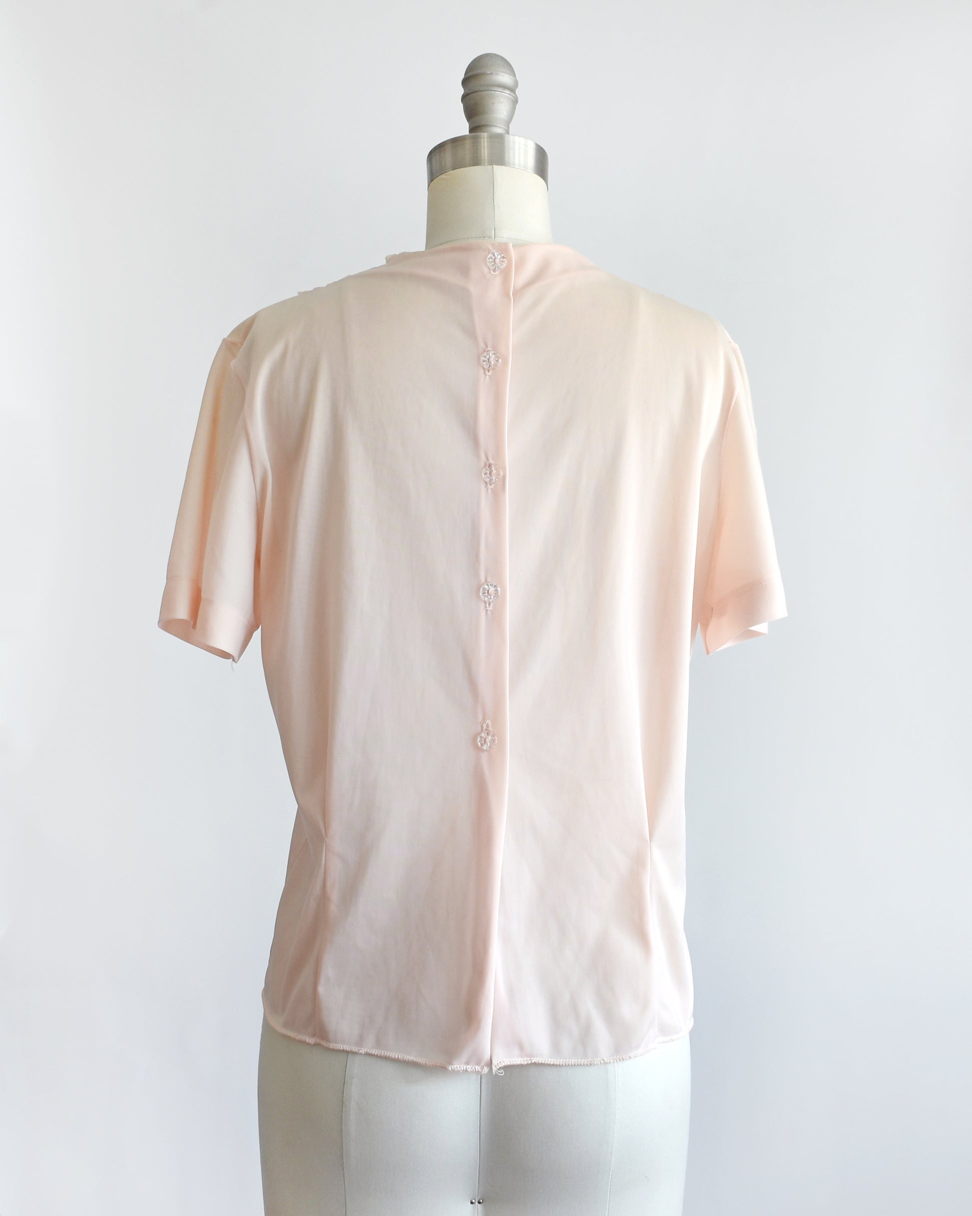 back view of a vintage 1960s pale blush pink short-sleeve blouse with a button up back, displayed on a white mannequin dress form against a white background.