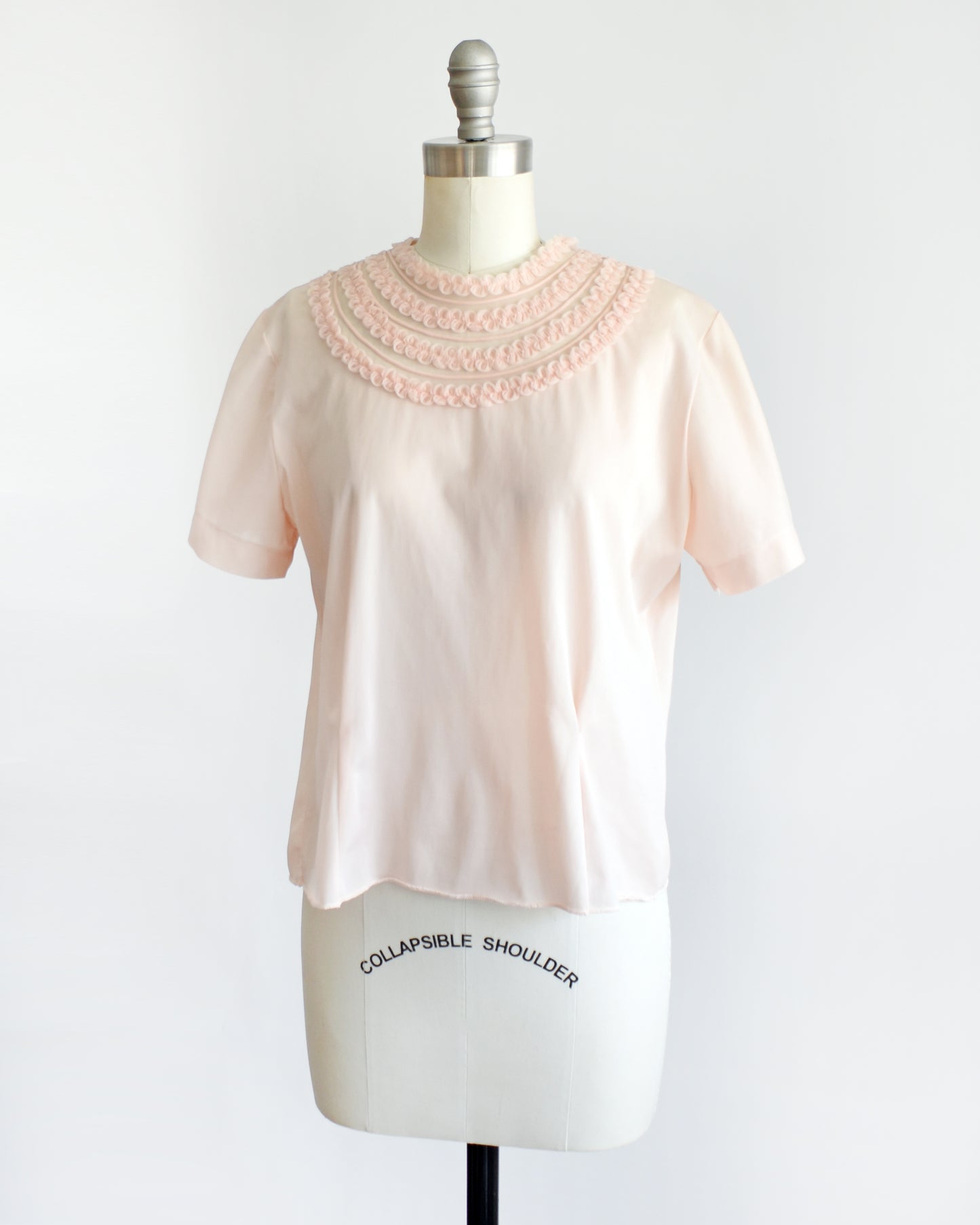 side front view of a vintage 1960s pale blush pink short-sleeve blouse with a decorative layered ruffle collar, displayed on a white mannequin dress form against a white background.
