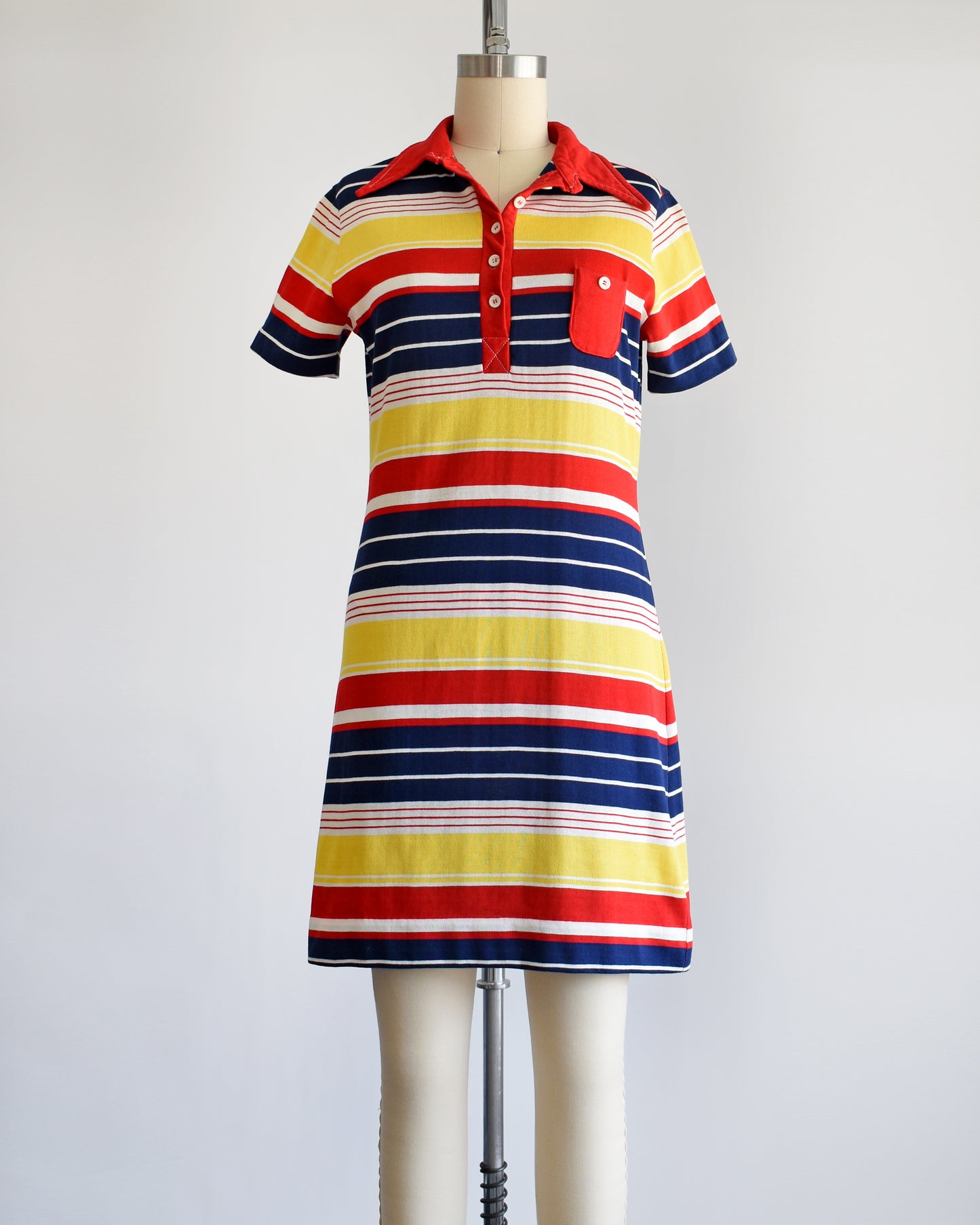 a vintage 1970s red, navy blue, yellow, and white striped dress with a red collar with six buttons down the front and a matching red chest pocket. three of the buttons are unbuttoned.