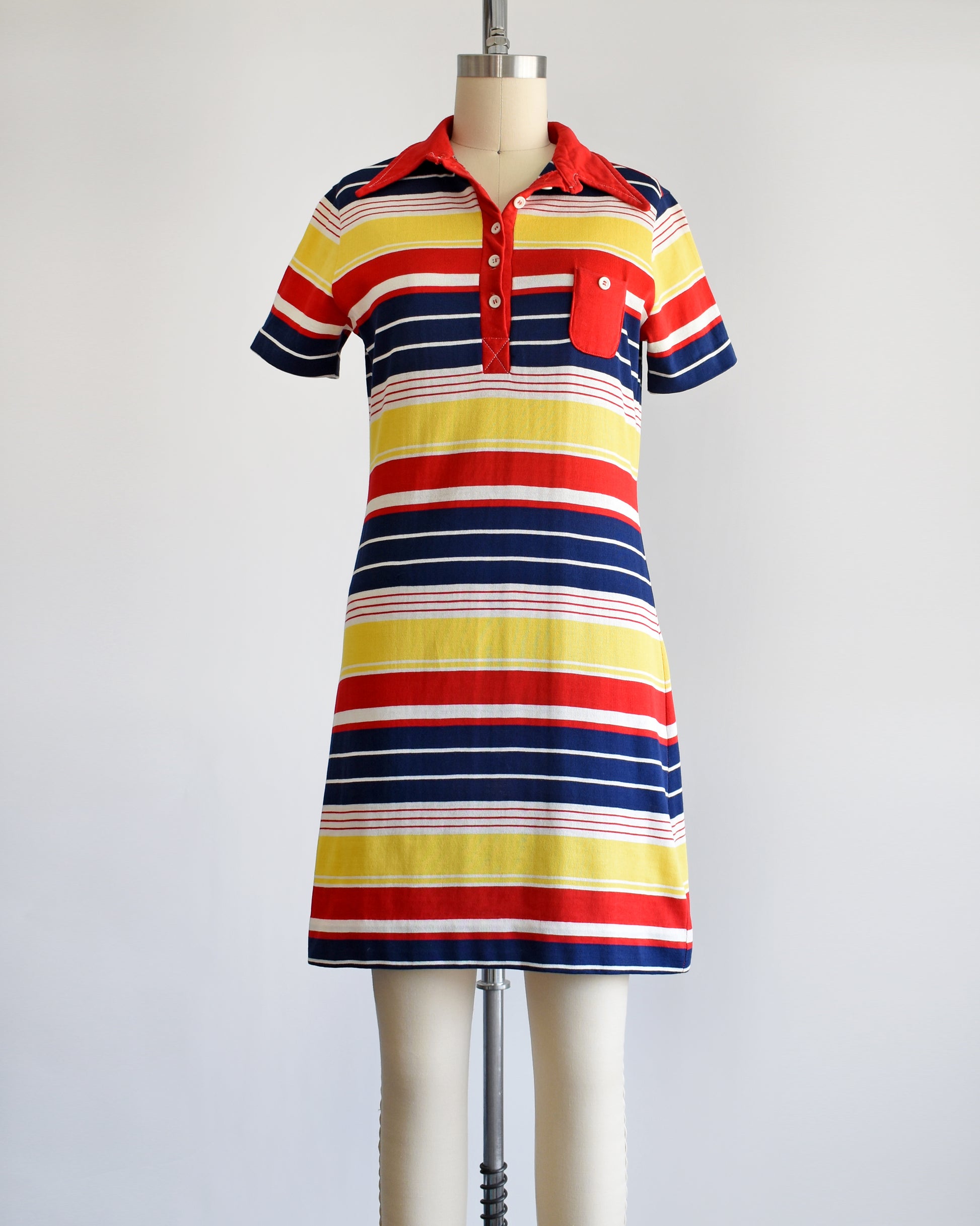 a vintage 1970s red, navy blue, yellow, and white striped dress with a red collar with six buttons down the front and a matching red chest pocket. three of the buttons are unbuttoned.