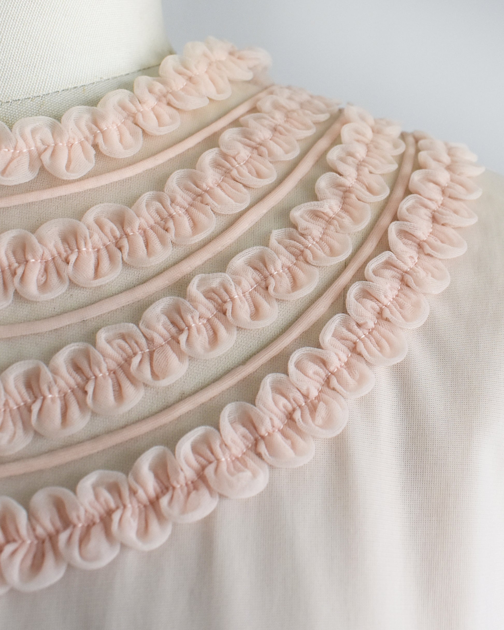 close up of the rows of ruffles around the collar on a vintage 1960s pale blush pink short-sleeve blouse with a decorative layered ruffle collar, displayed on a white mannequin dress form against a white background.