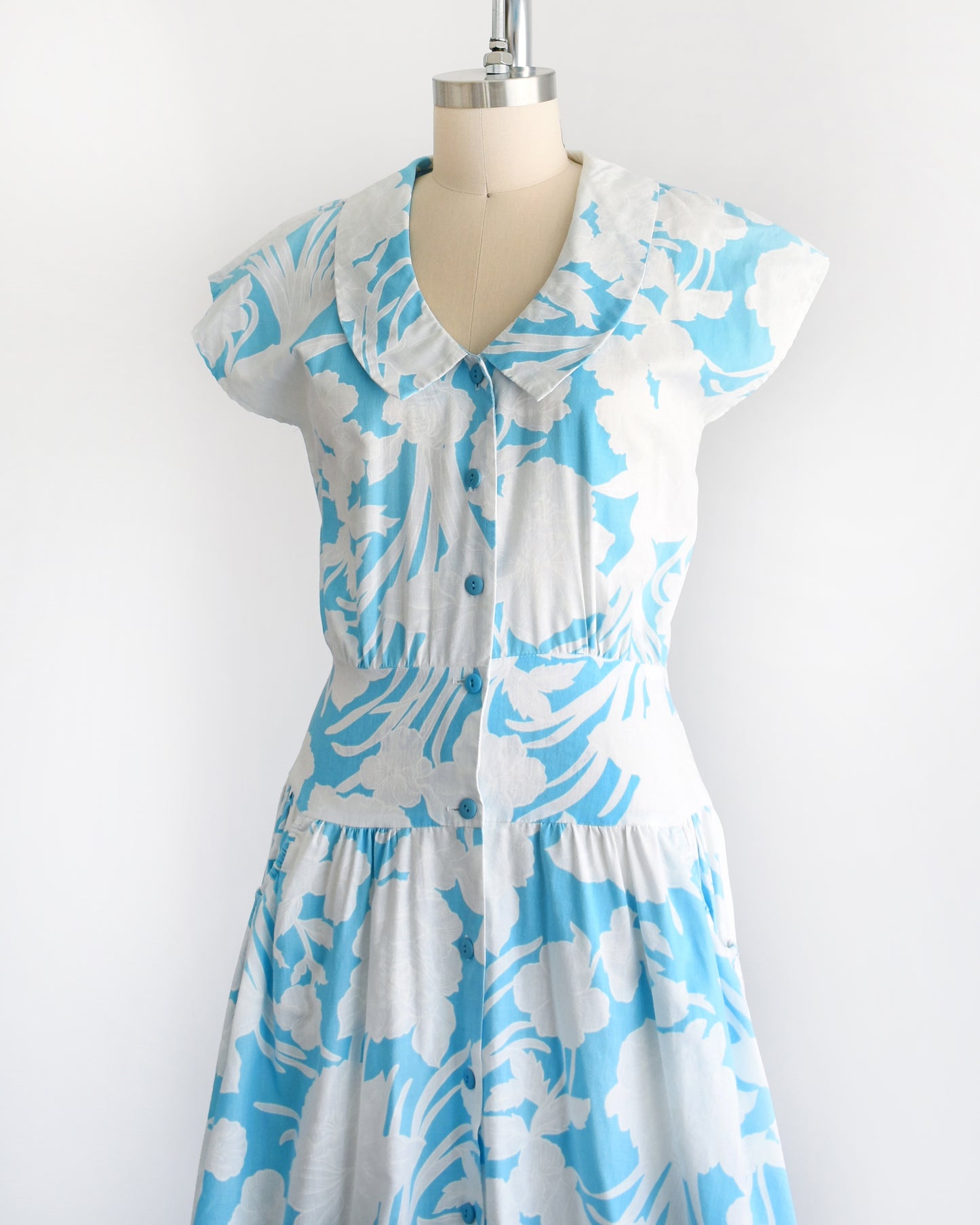 Side front view of a vintage 80s floral dress that is light blue with white floral print. Peter Pan style collar with blue buttons down the front. Cap sleeves. Wide waistband and drop waist skirt.