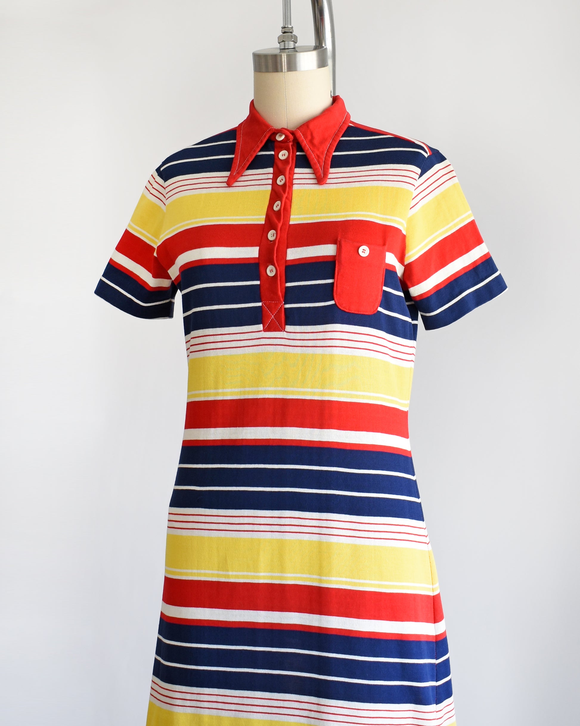 a vintage 1970s red, navy blue, yellow, and white striped dress with a red collar with six buttons down the front and a matching red chest pocket. the buttons are all buttoned.