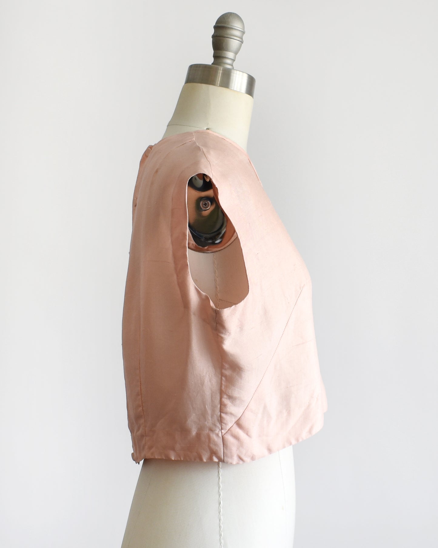 side view of a vintage 1950s/1960s pale pink sleeveless silk crop top displayed on a white dress form mannequin against a plain white background. 