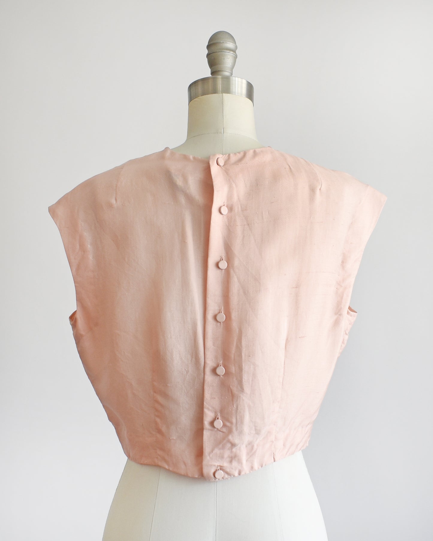 back view of a vintage 1950s/1960s pale pink sleeveless silk crop top with button back displayed on a white dress form mannequin against a plain white background. 