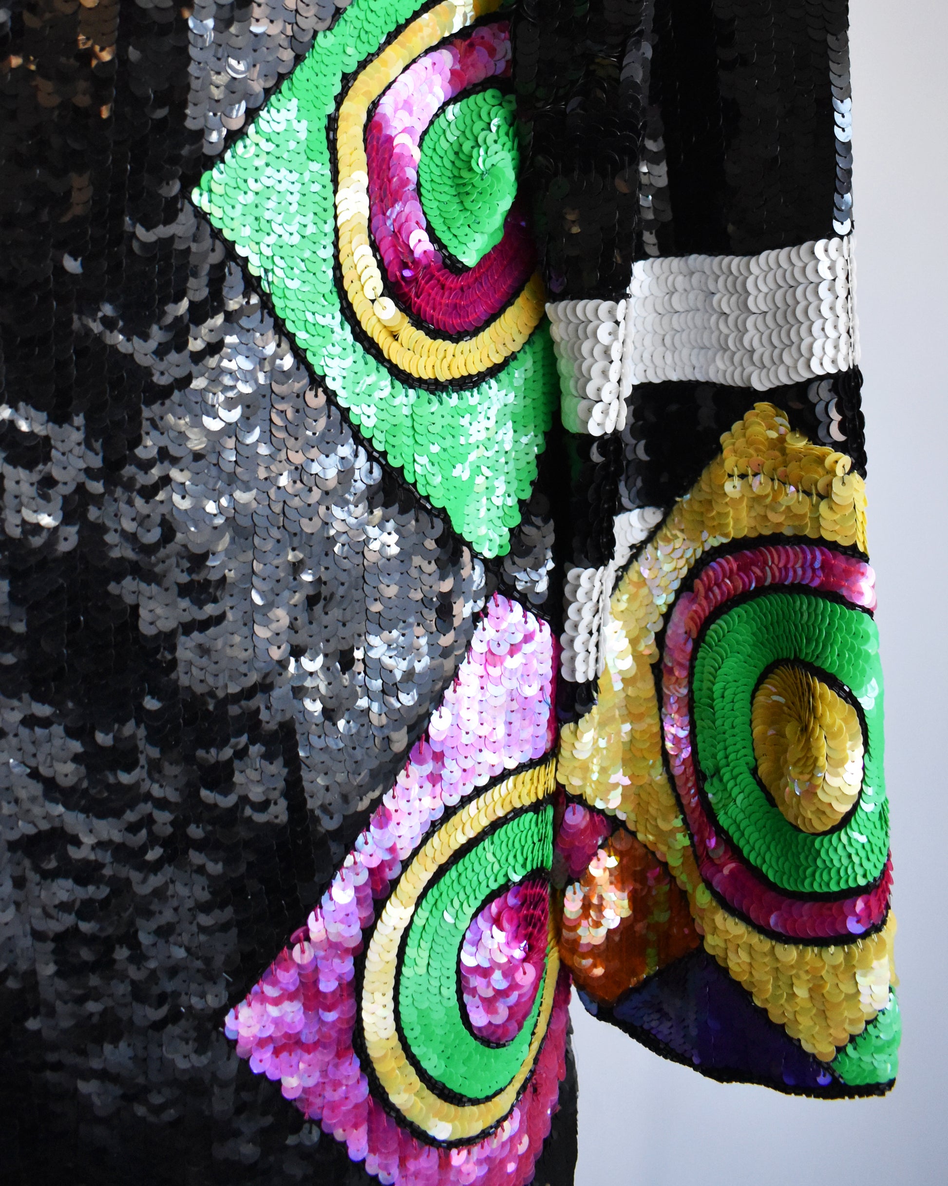 close up of the sleeves and side of  a vintage 1980s 1990s black sequin cardigan jacket with colorful abstract patterns on a mannequin against a white background