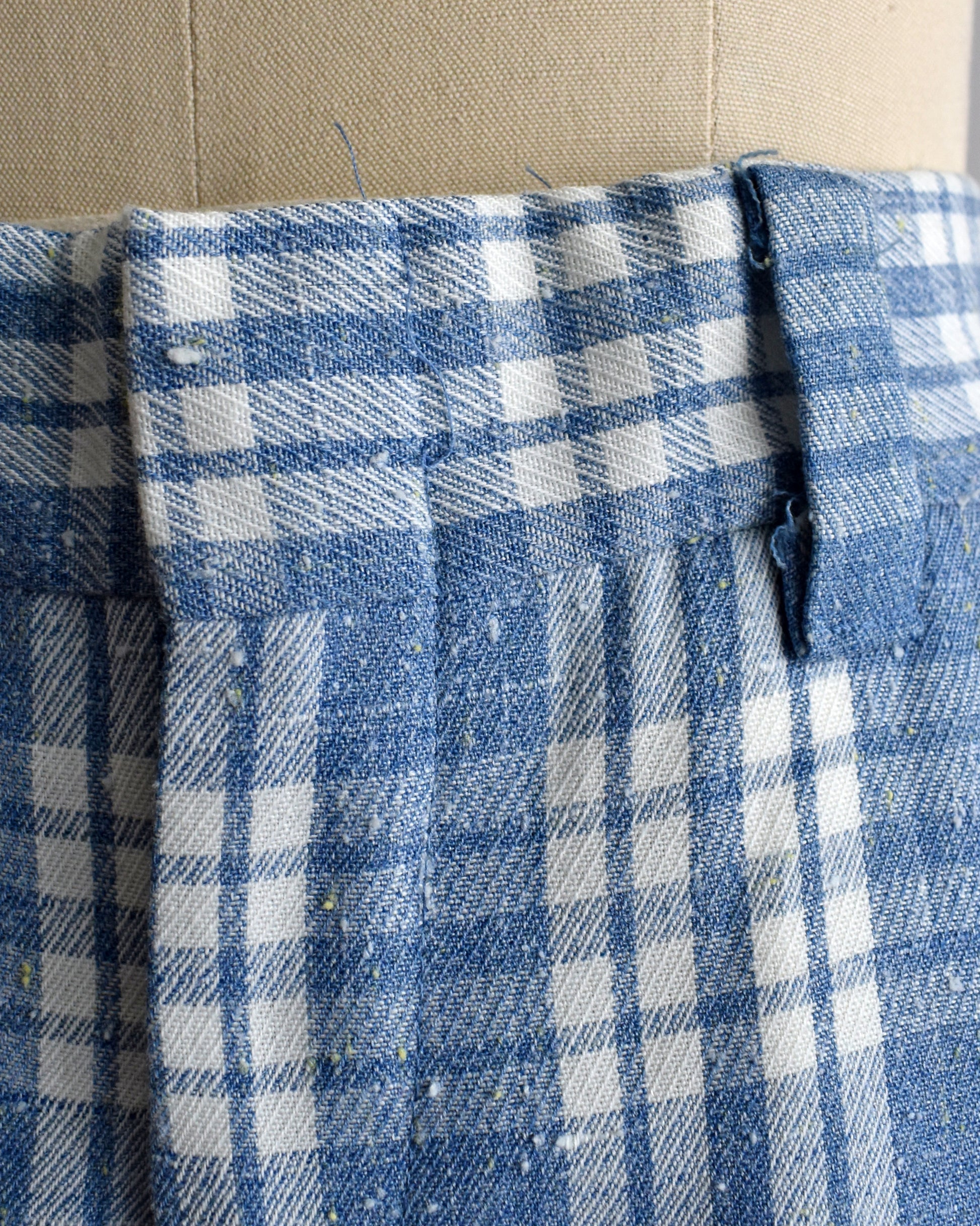 Close-up of a blue and white plaid pants, shown on a beige dress form with a waistband and button detail visible.