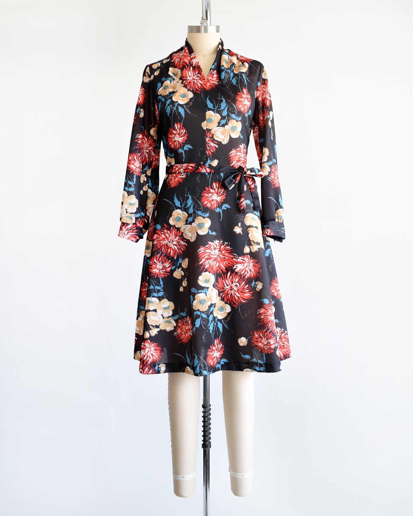 A vintage black dress that has a large red, tan, white, and blue floral print, a v-neckline, long sleeves, and matching tie belt.