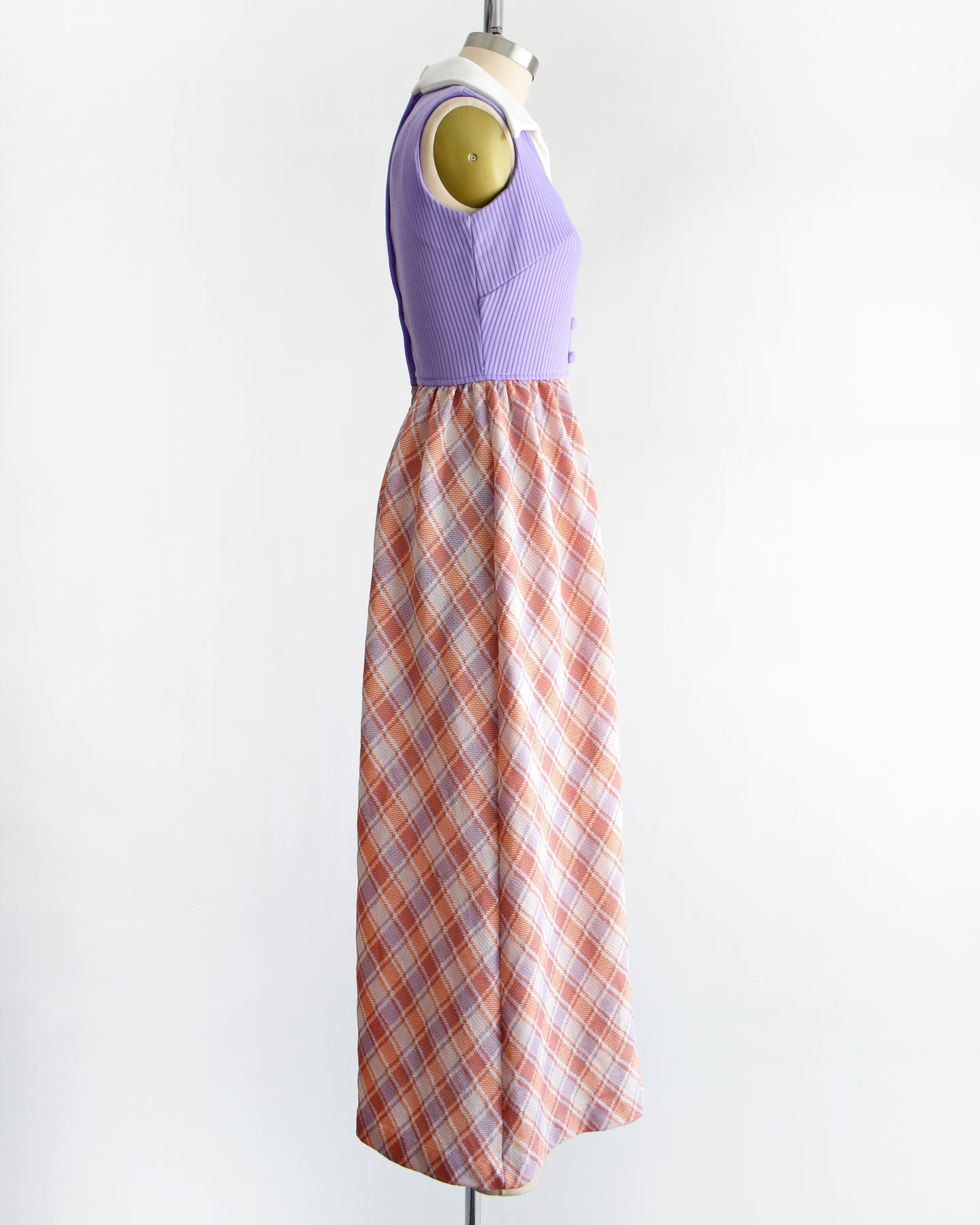 side view of a vintage 1970s sleeveless maxi dress displayed on a mannequin against a plain white background. The dress features a lavender purple ribbed bodice with a white collar and button details, paired with a pleated skirt in a peach, orange, and white plaid pattern.