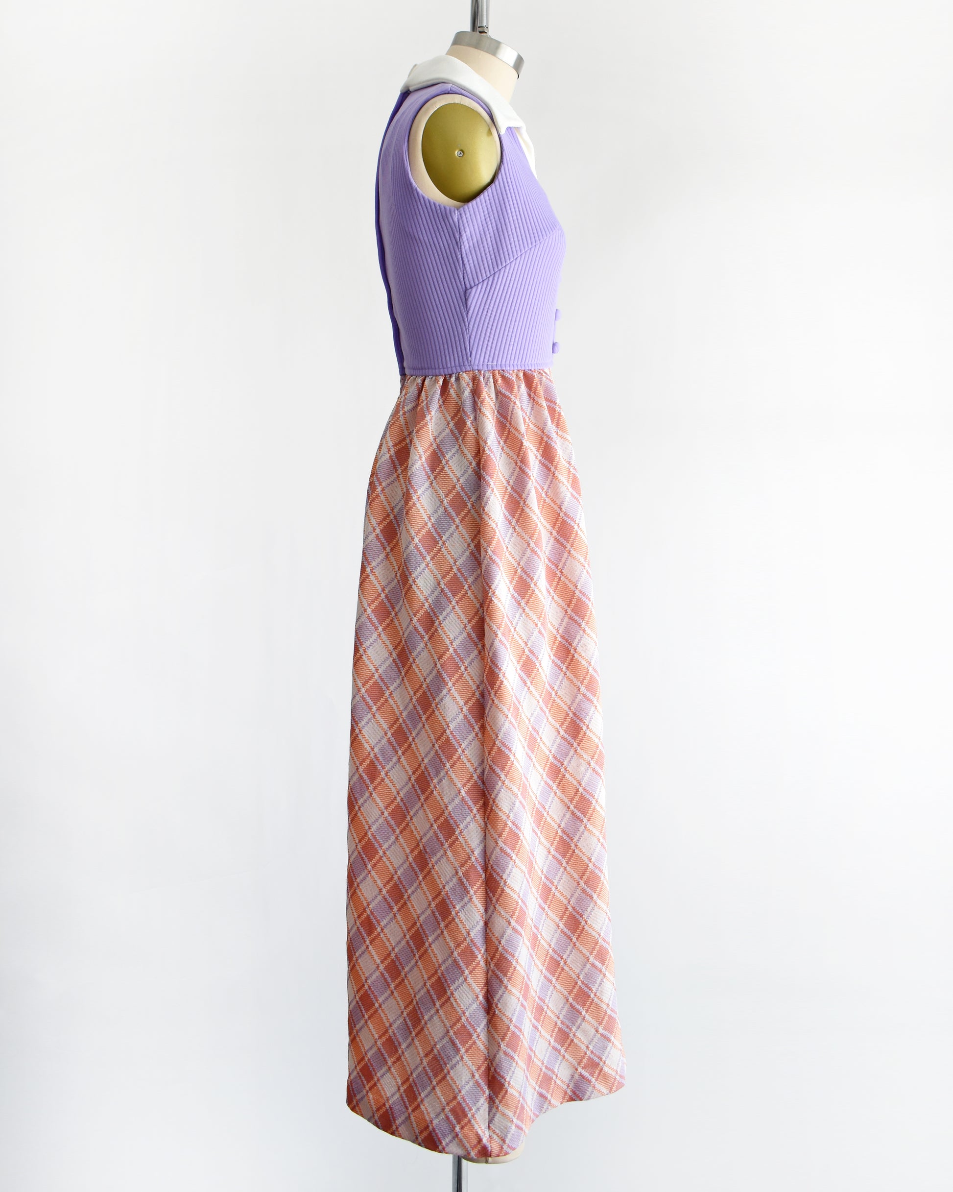 side view of a vintage 1970s sleeveless maxi dress displayed on a mannequin against a plain white background. The dress features a lavender purple ribbed bodice with a white collar and button details, paired with a pleated skirt in a peach, orange, and white plaid pattern.