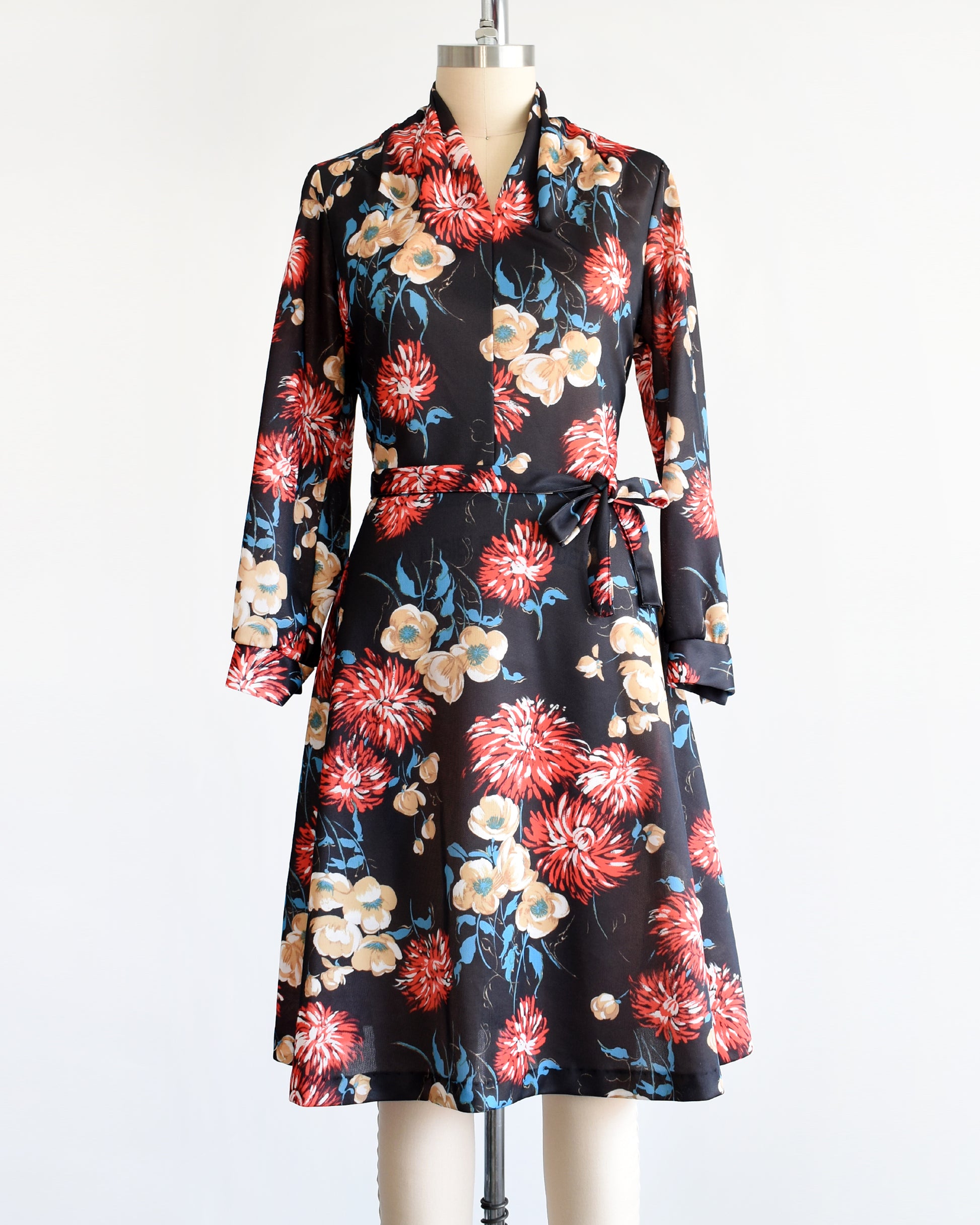 A vintage black dress that has a large red, tan, white, and blue floral print, a v-neckline, long sleeves, and matching tie belt.