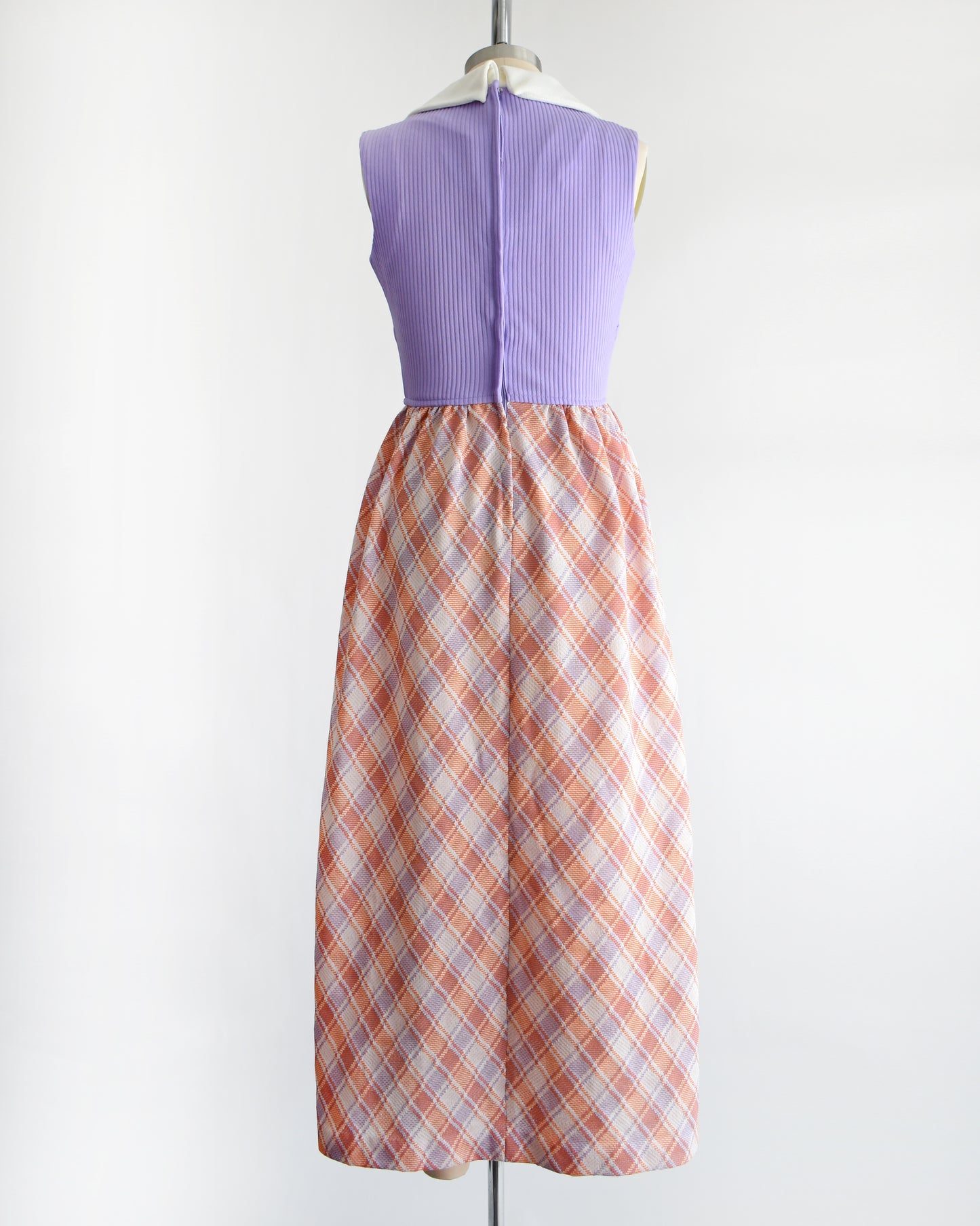 back view of a vintage 1970s sleeveless maxi dress displayed on a mannequin against a plain white background. The dress features a lavender purple ribbed bodice with a white collar and button details, paired with a pleated skirt in a peach, orange, and white plaid pattern.
