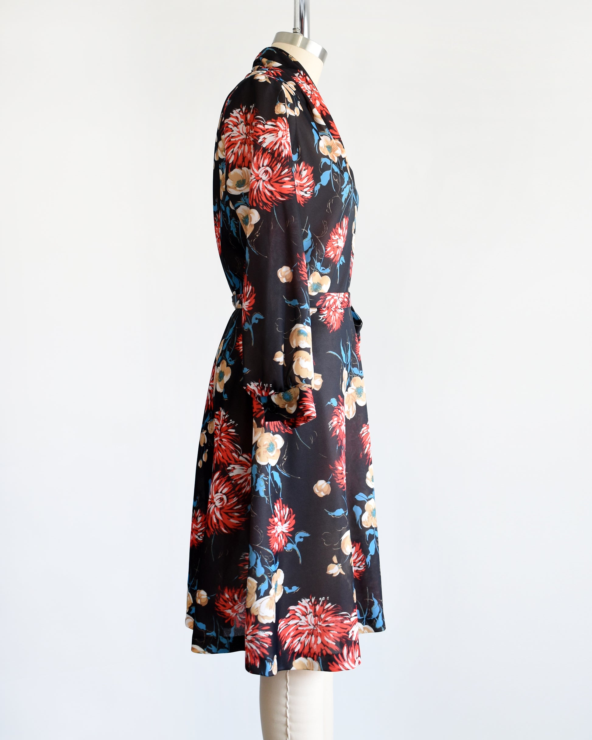 side view of a vintage black dress that has a large red, tan, white, and blue floral print, a v-neckline, long sleeves, and matching tie belt.