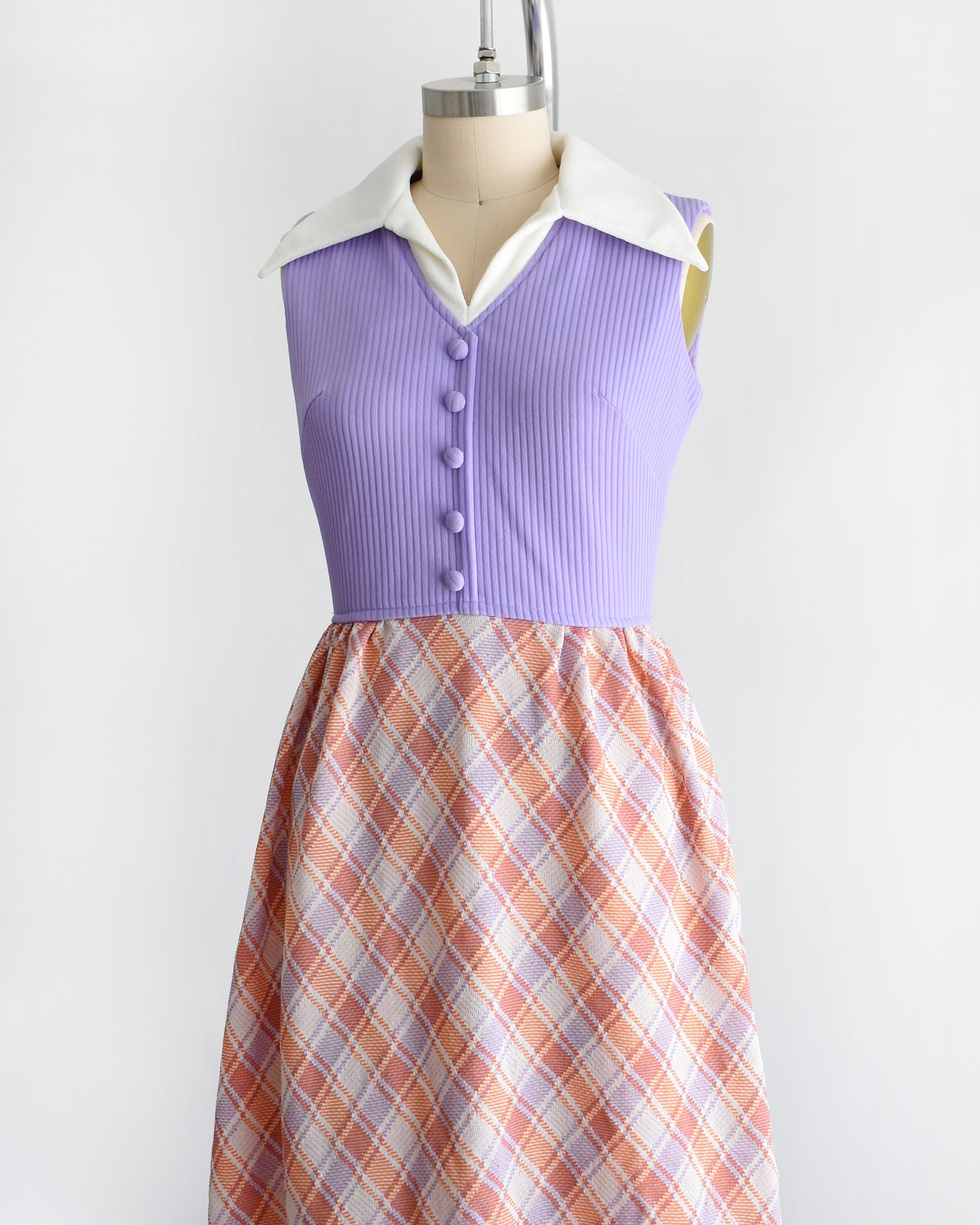 side front view of a vintage 1970s sleeveless maxi dress displayed on a mannequin against a plain white background. The dress features a lavender purple ribbed bodice with a white collar and button details, paired with a pleated skirt in a peach, orange, and white plaid pattern.