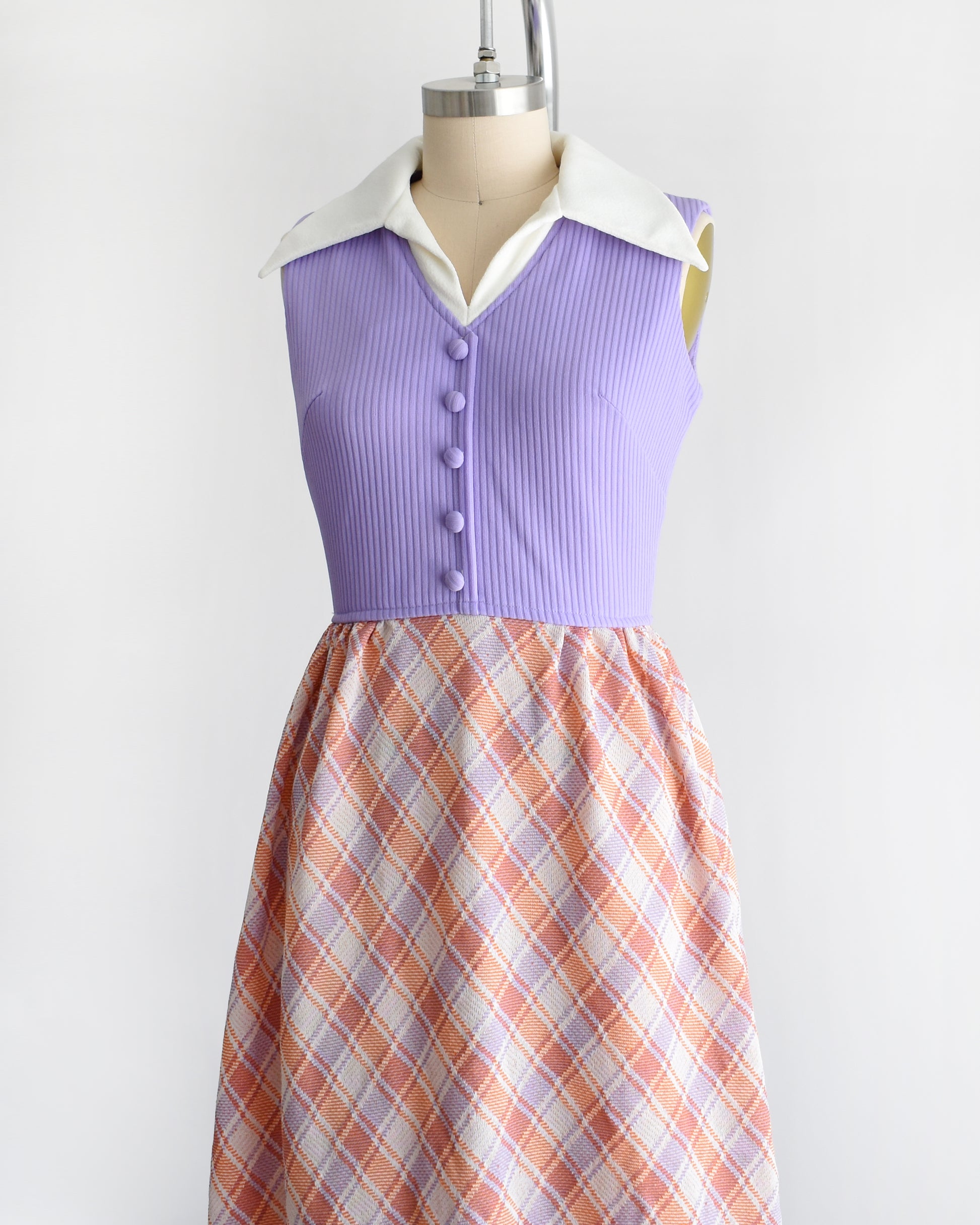 side front view of a vintage 1970s sleeveless maxi dress displayed on a mannequin against a plain white background. The dress features a lavender purple ribbed bodice with a white collar and button details, paired with a pleated skirt in a peach, orange, and white plaid pattern.