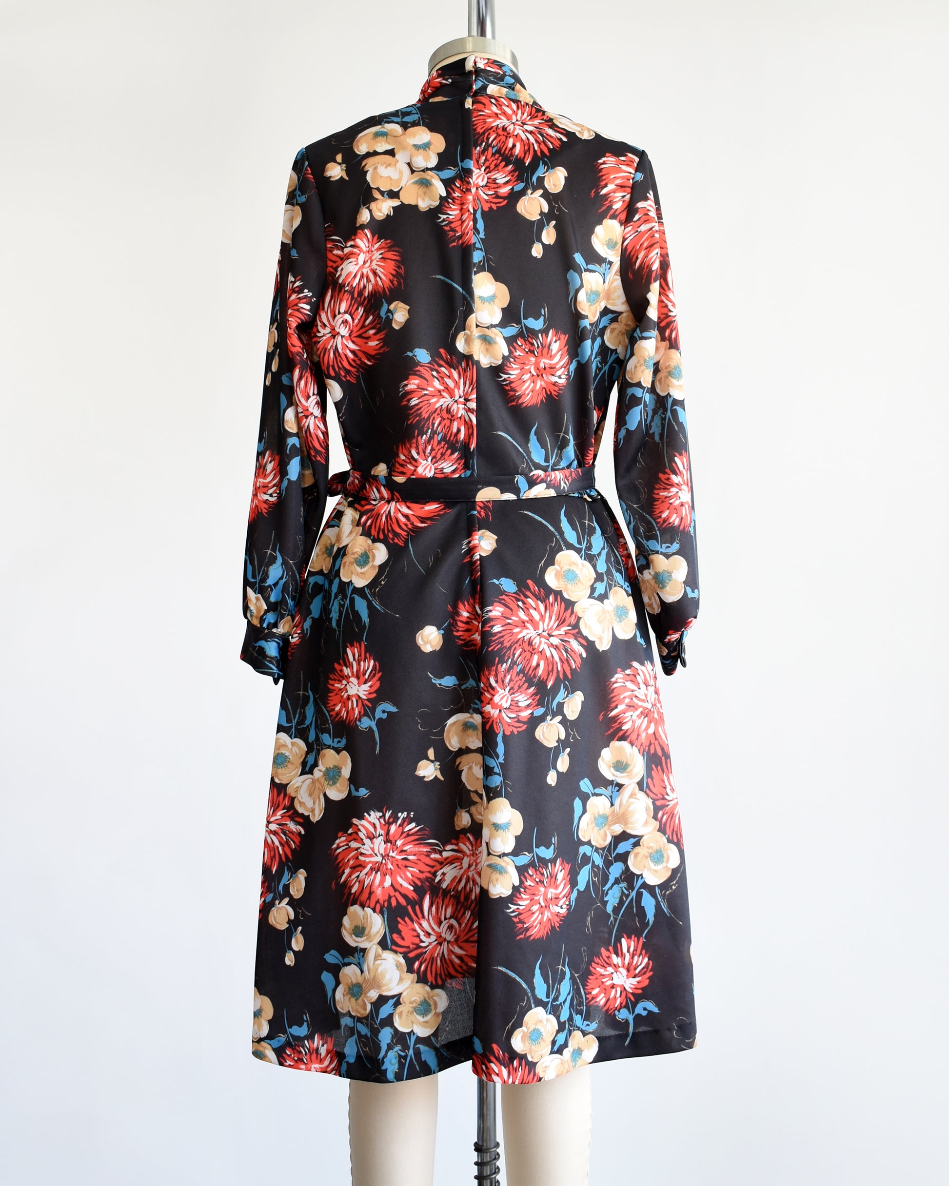 back view of a vintage black dress that has a large red, tan, white, and blue floral print, long sleeves, and matching tie belt.