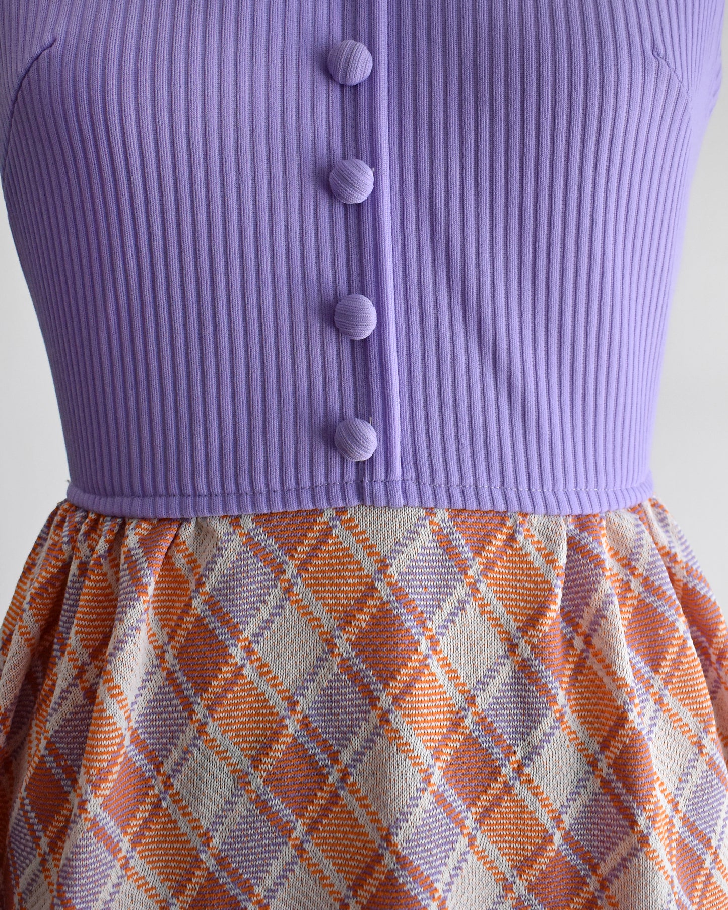 close up of the bodice and waist on a vintage 1970s sleeveless maxi dress displayed on a mannequin against a plain white background. The dress features a lavender purple ribbed bodice with a white collar and button details, paired with a pleated skirt in a peach, orange, and white plaid pattern.