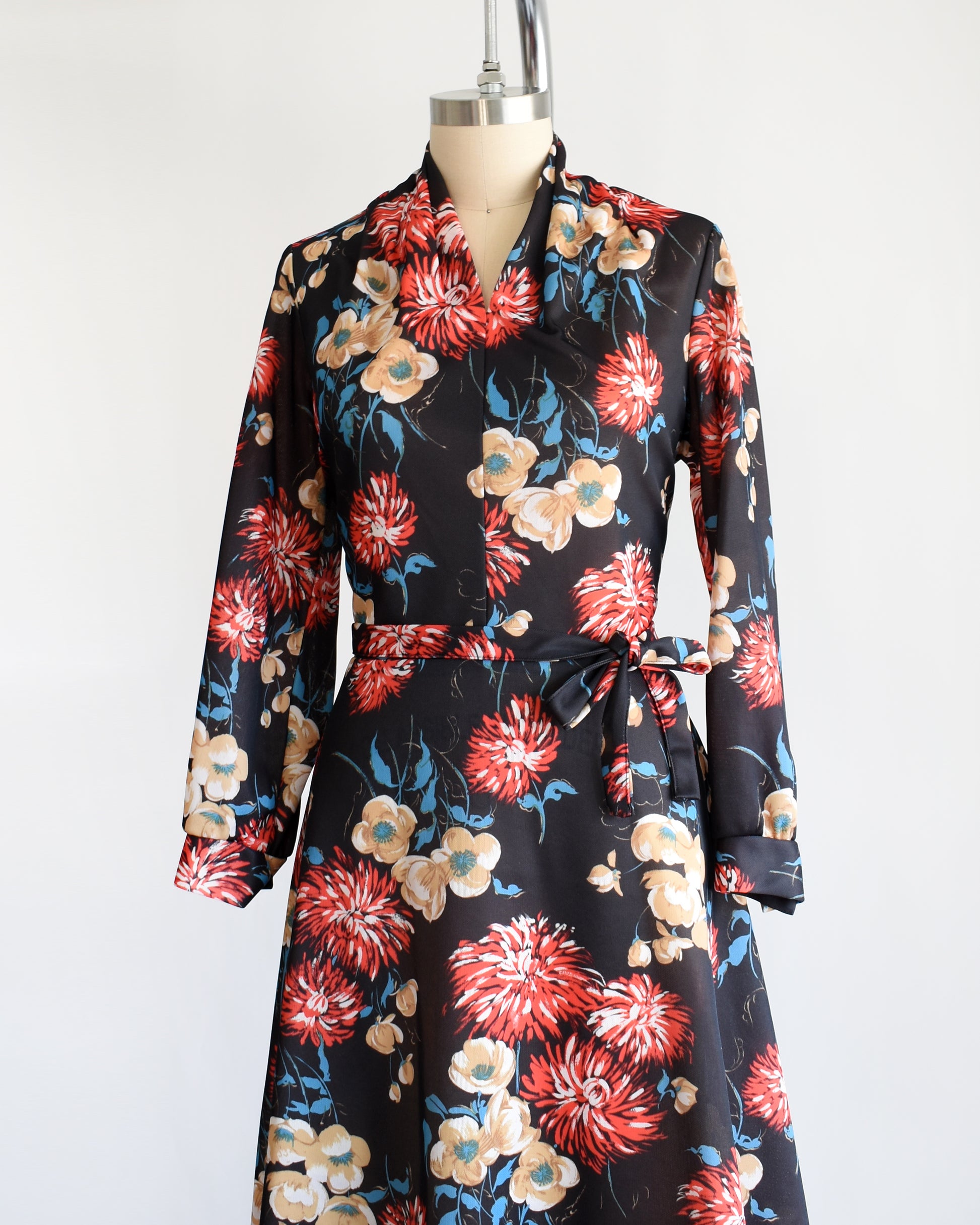 side front view of a vintage black dress that has a large red, tan, white, and blue floral print, a v-neckline, long sleeves, and matching tie belt.