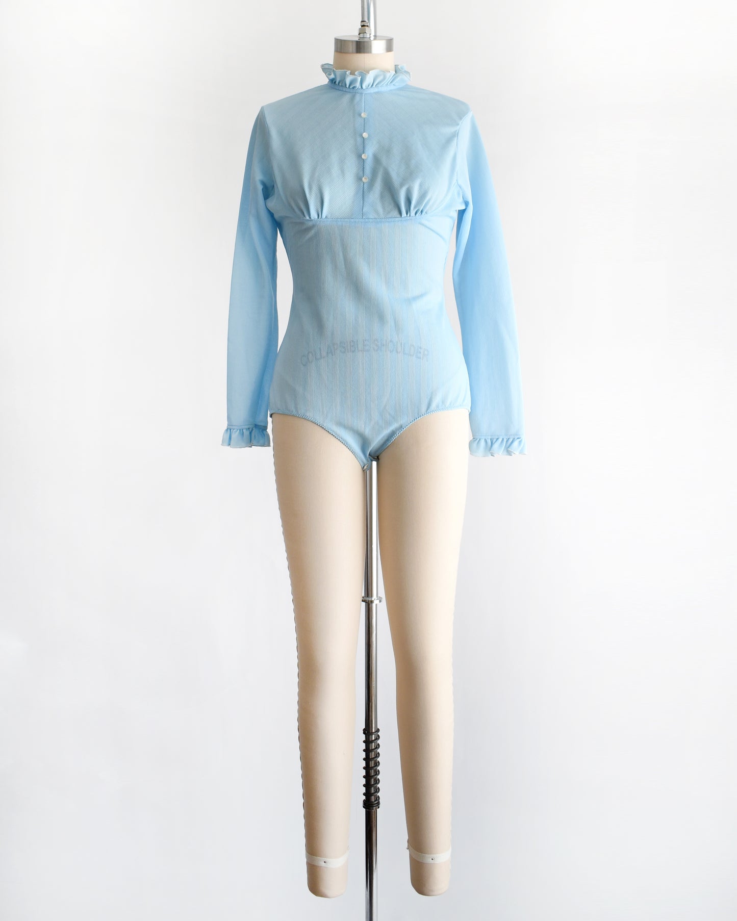 A vintage 1970s light blue long-sleeve bodysuit with a ruffled mock neck and button front displayed on a beige mannequin against a plain white background.