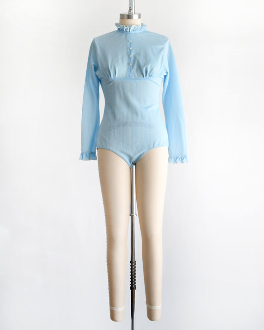 A vintage 1970s light blue long-sleeve bodysuit with a ruffled mock neck and button front displayed on a beige mannequin against a plain white background.