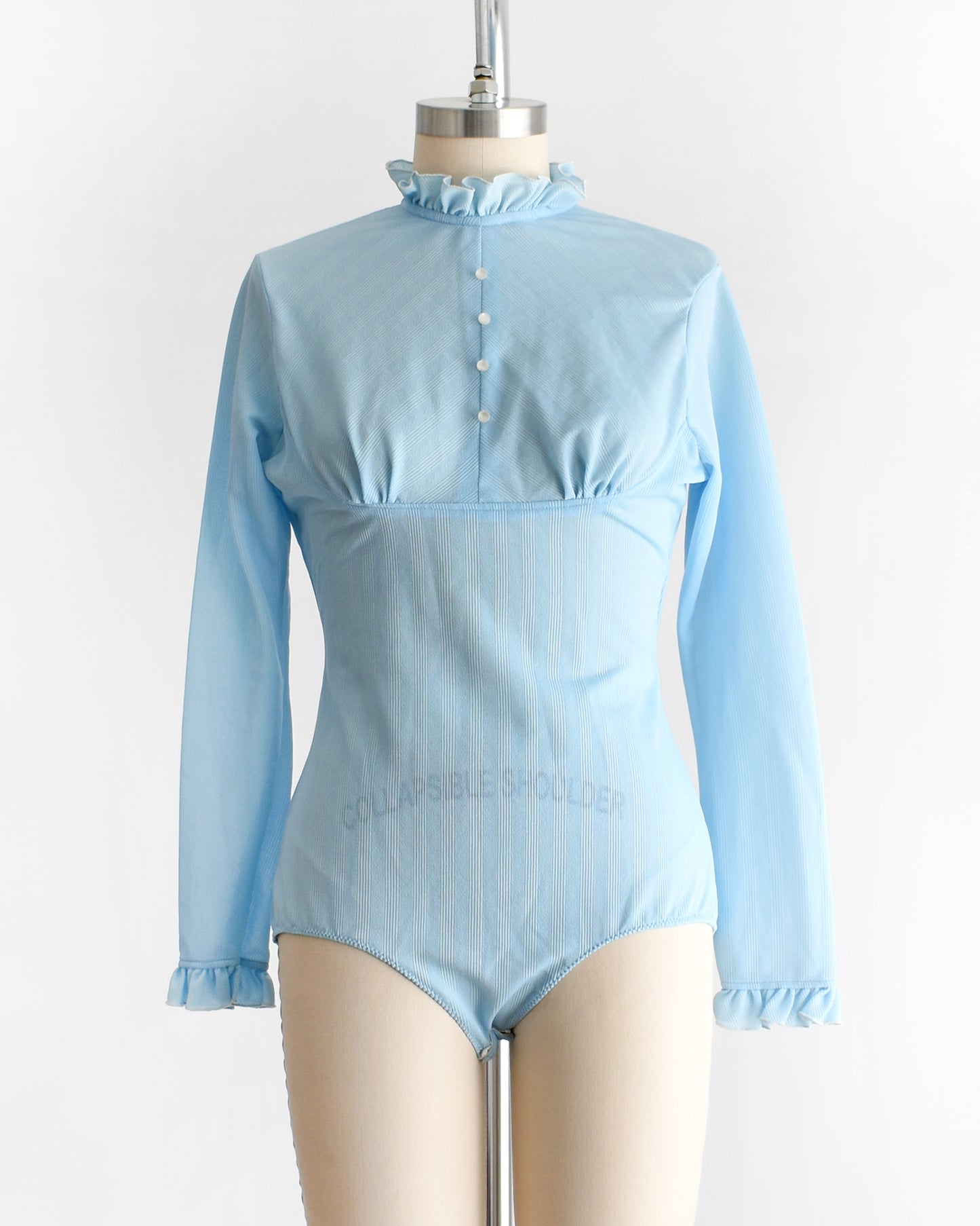 A vintage 1970s light blue long-sleeve bodysuit with a ruffled mock neck and button front displayed on a beige mannequin against a plain white background.