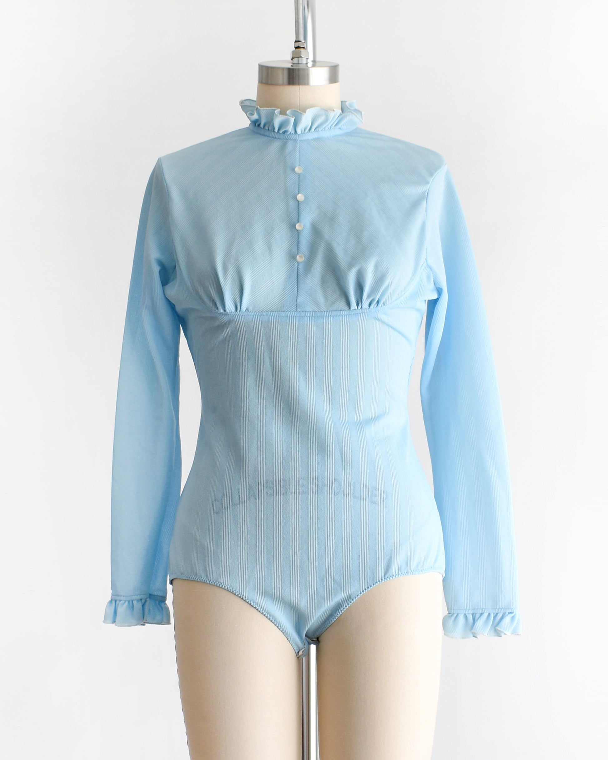 A vintage 1970s light blue long-sleeve bodysuit with a ruffled mock neck and button front displayed on a beige mannequin against a plain white background.