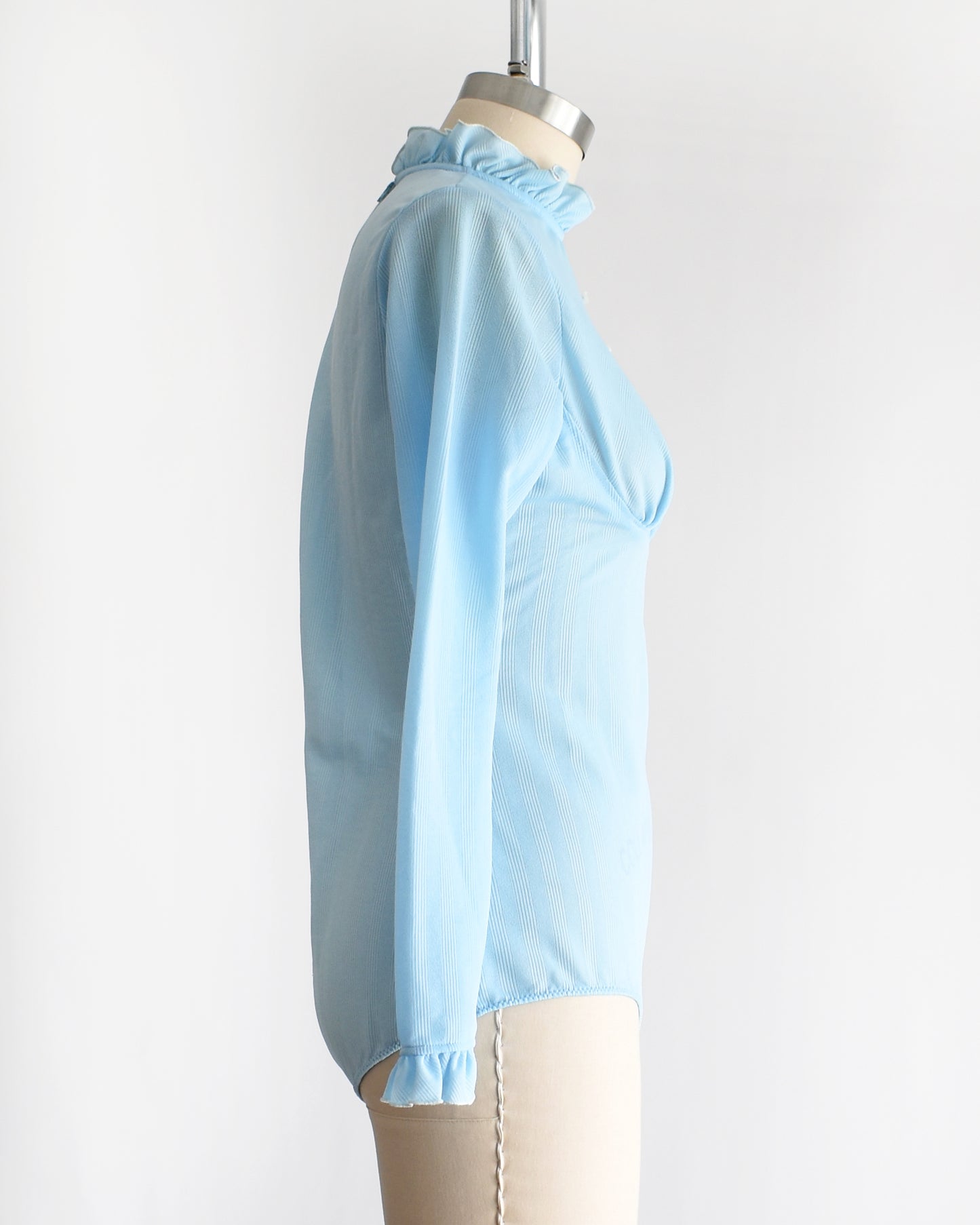 side view of a vintage 1970s light blue long-sleeve bodysuit with a ruffled mock neck and button front displayed on a beige mannequin against a plain white background.