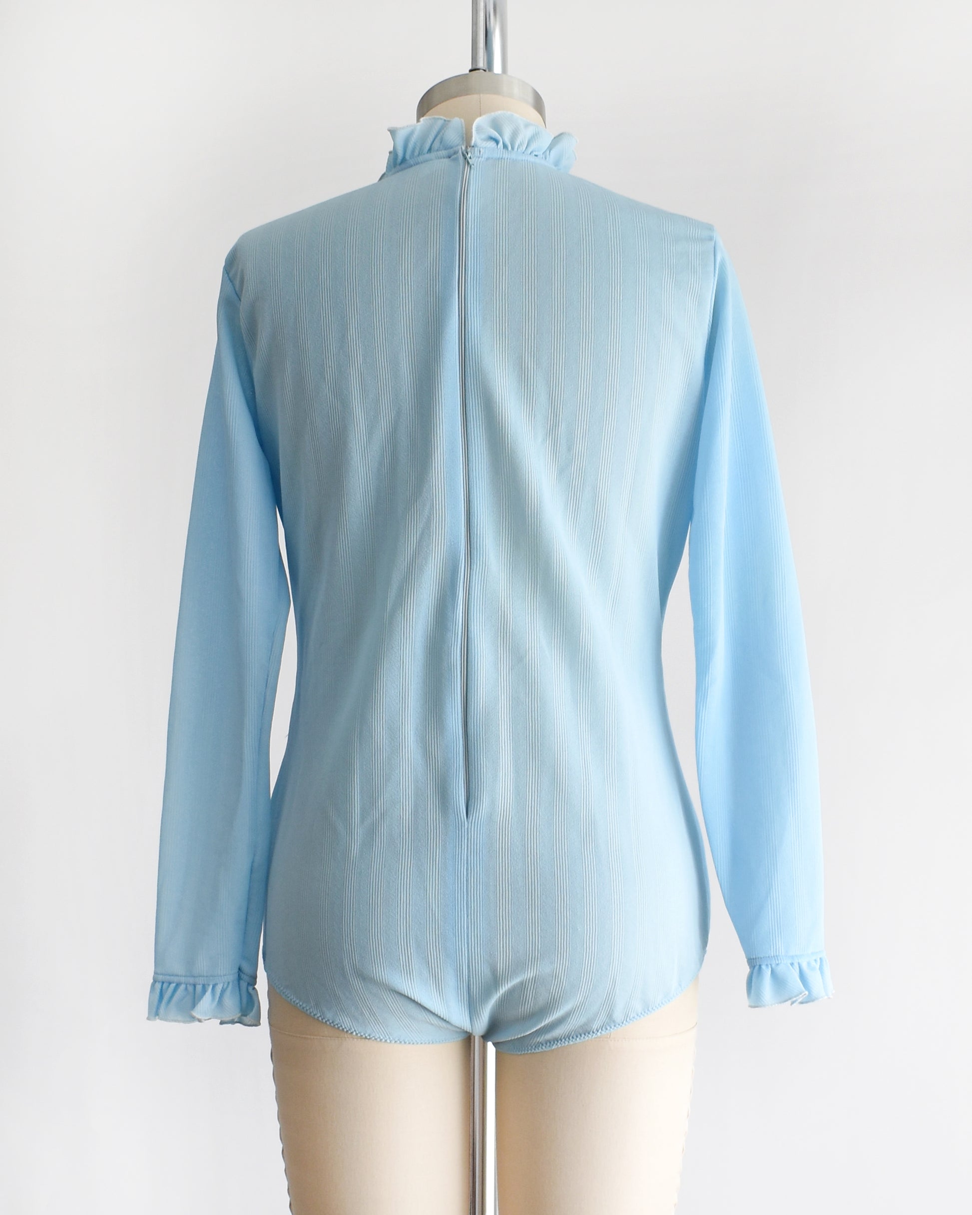back view of a vintage 1970s light blue long-sleeve bodysuit with a ruffled mock neck and button front displayed on a beige mannequin against a plain white background.