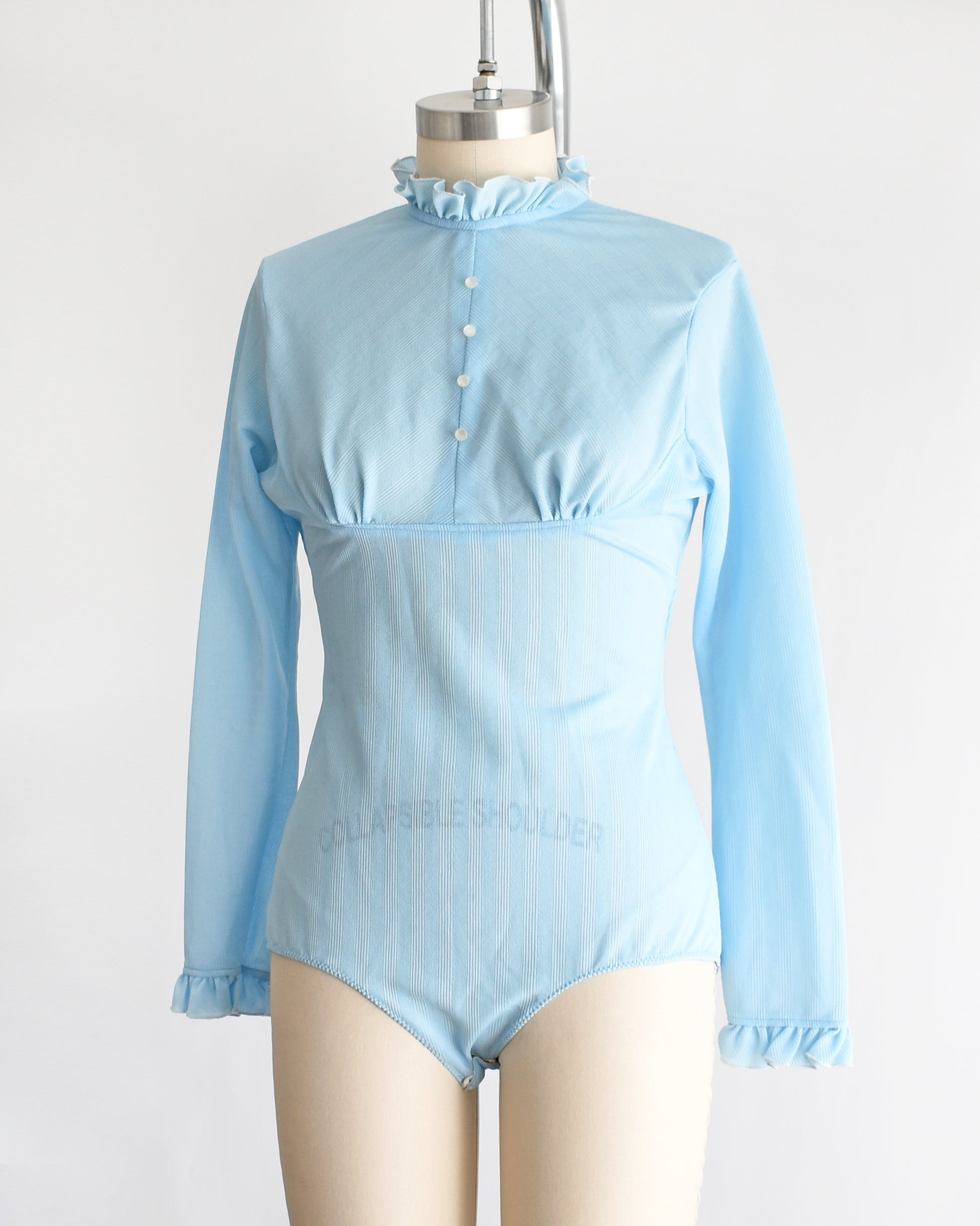 side front view of a vintage 1970s light blue long-sleeve bodysuit with a ruffled mock neck and button front displayed on a beige mannequin against a plain white background.