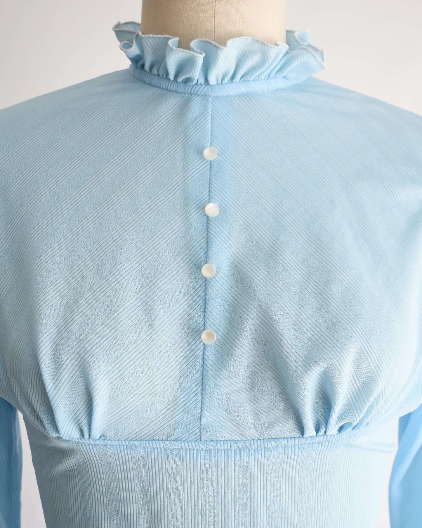 close up of the bodice on a vintage 1970s light blue long-sleeve bodysuit with a ruffled mock neck and button front displayed on a beige mannequin against a plain white background.