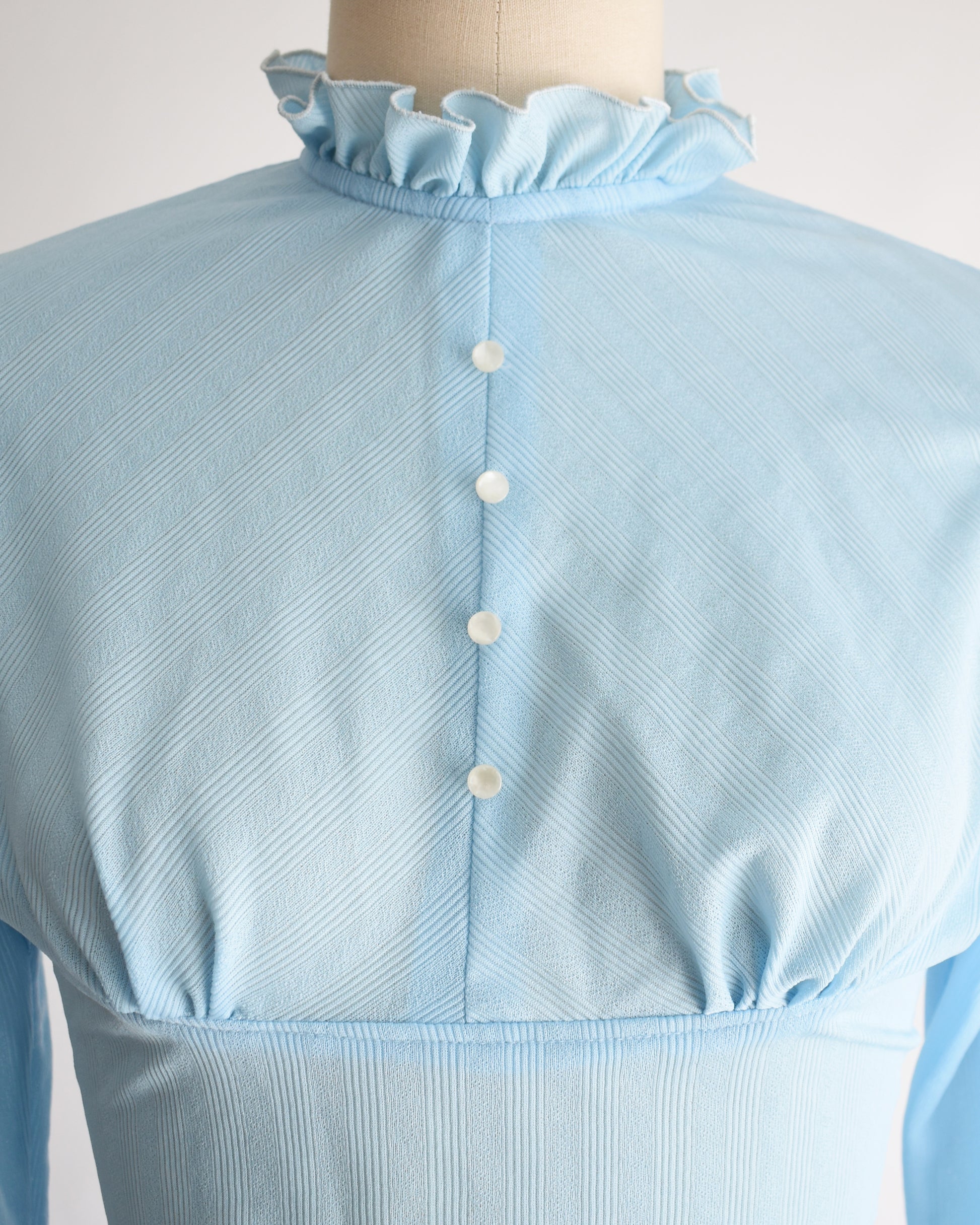 close up of the bodice on a vintage 1970s light blue long-sleeve bodysuit with a ruffled mock neck and button front displayed on a beige mannequin against a plain white background.
