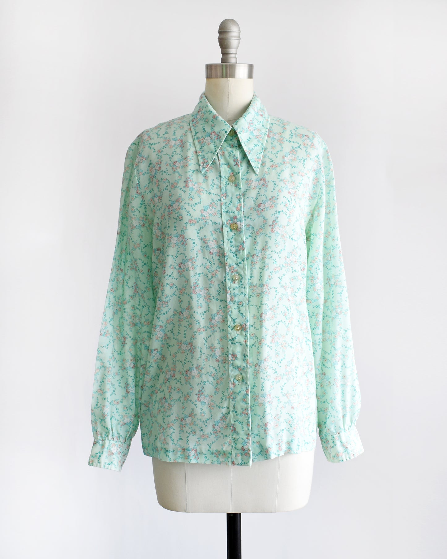 a vintage 1970s long-sleeve button-up blouse with a light mint green and pink floral pattern, displayed on a cream dress form mannequin against a white background.