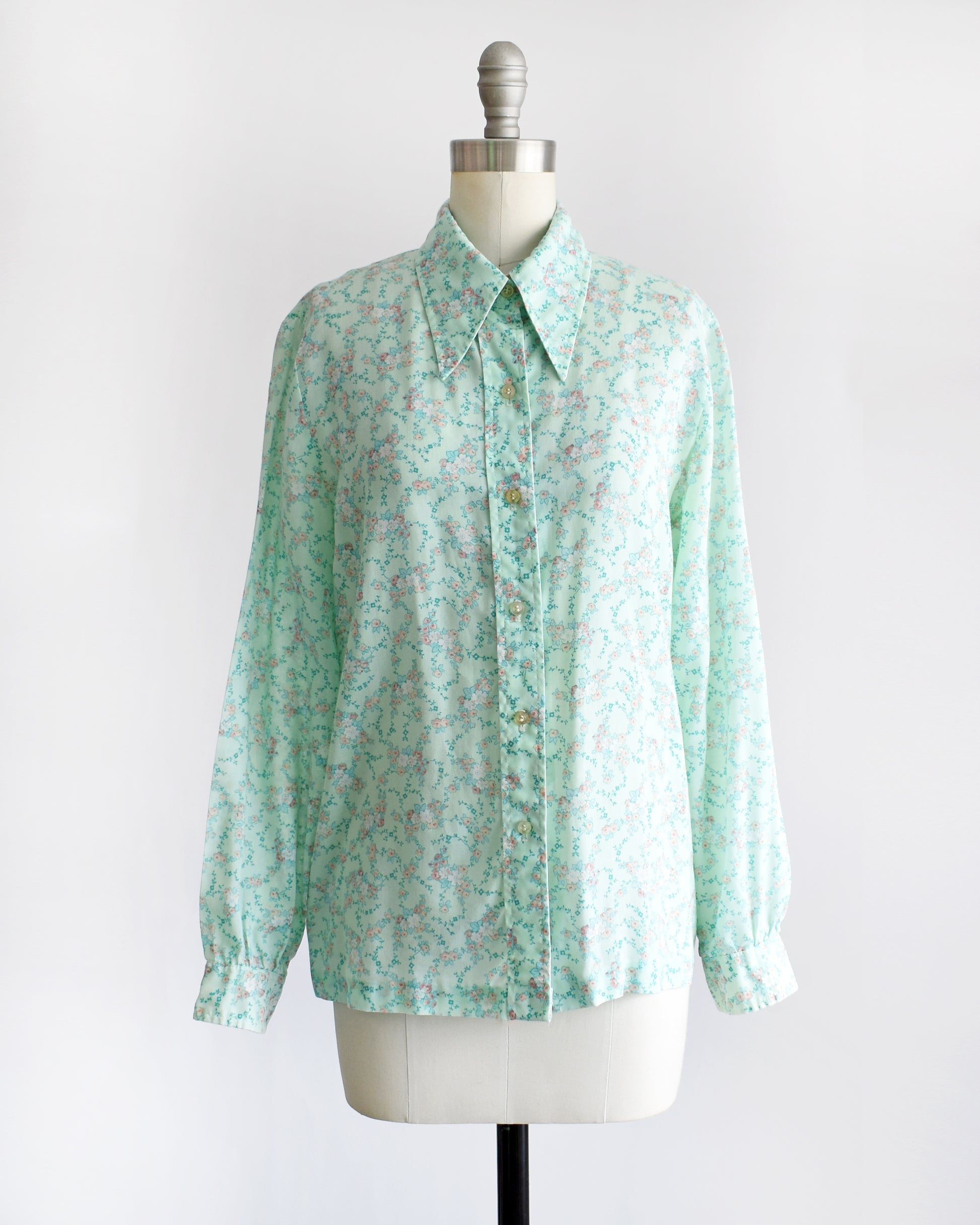 a vintage 1970s long-sleeve button-up blouse with a light mint green and pink floral pattern, displayed on a cream dress form mannequin against a white background.