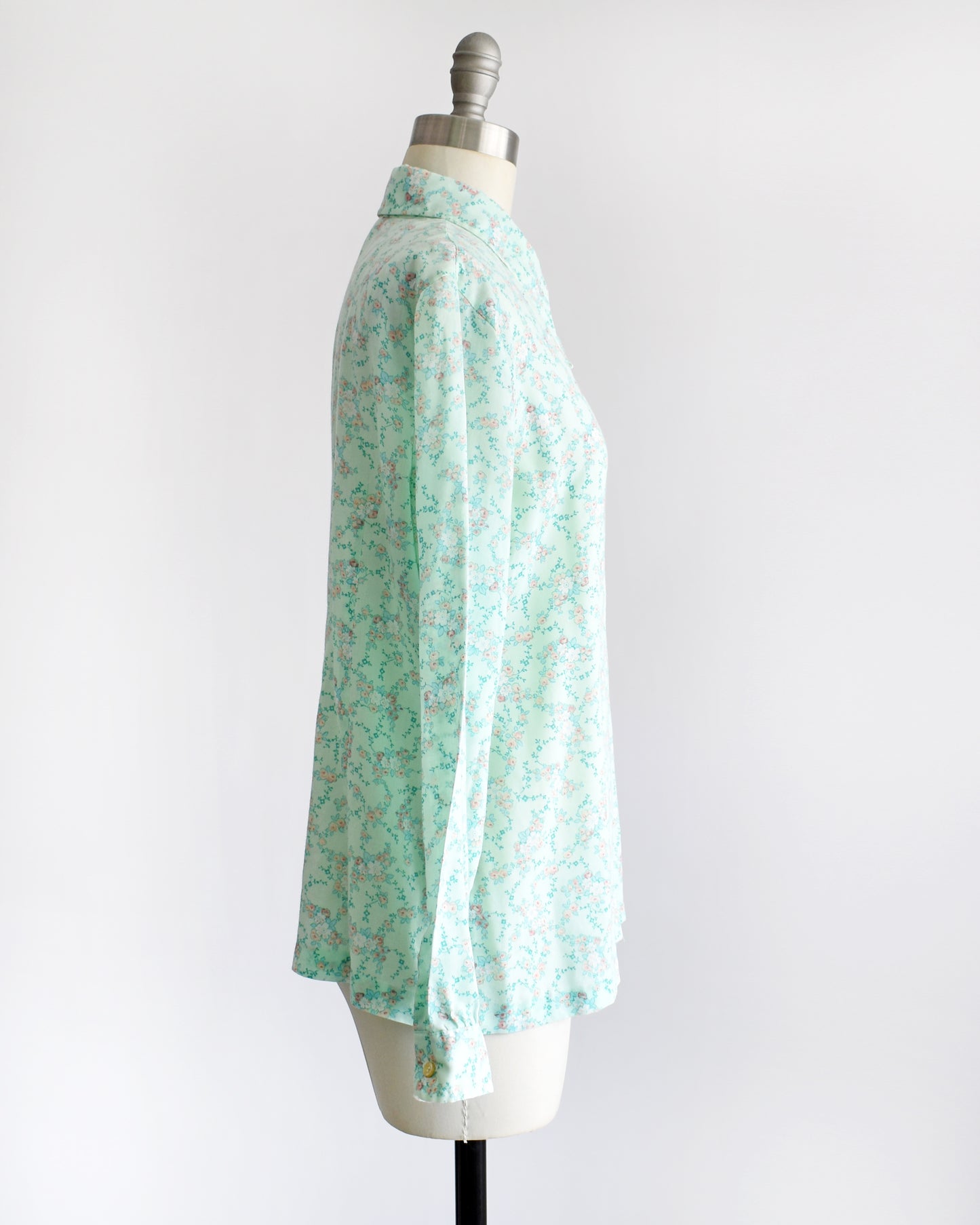 side view of a vintage 1970s long-sleeve button-up blouse with a light mint green and pink floral pattern, displayed on a cream dress form mannequin against a white background.