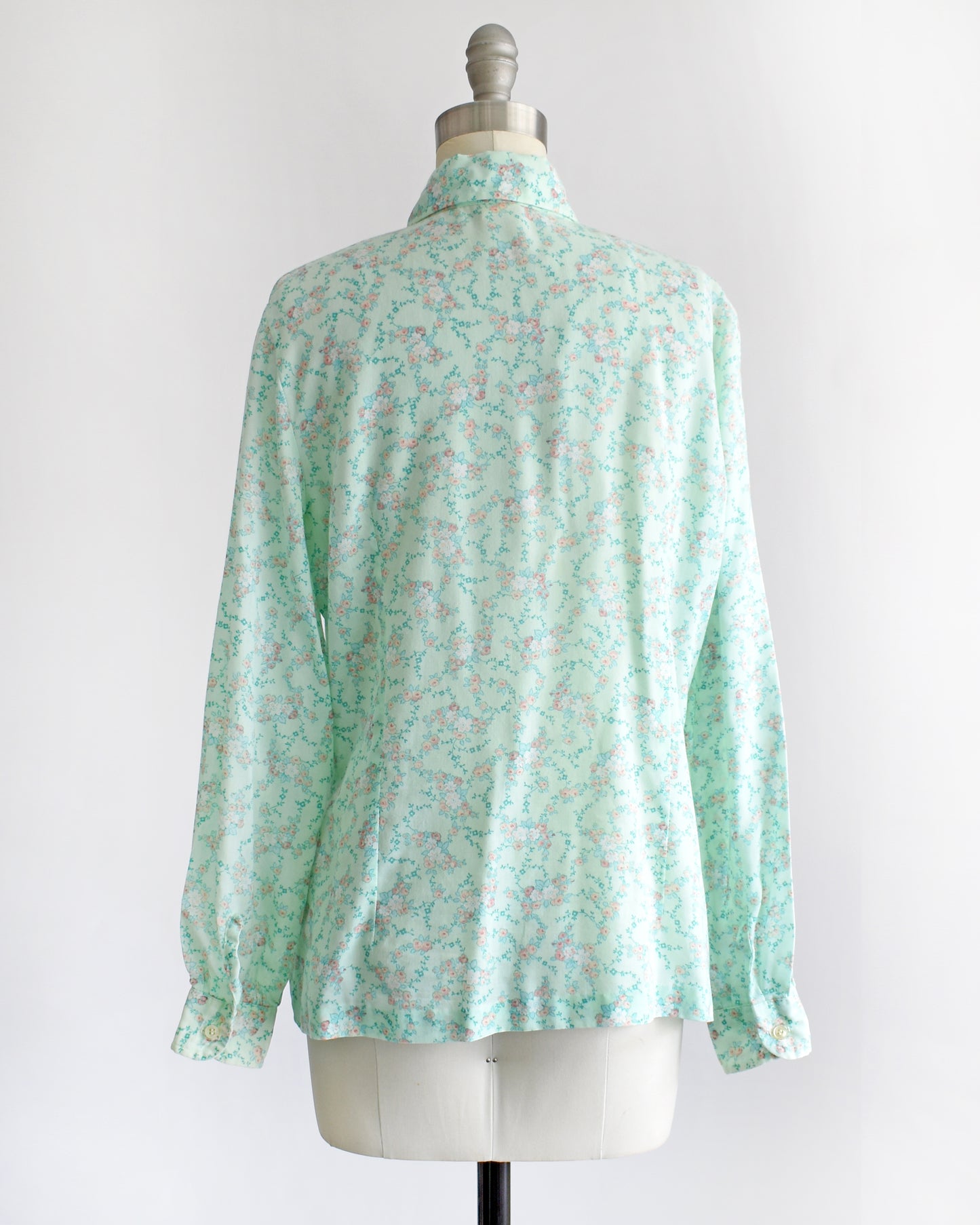 back view of a vintage 1970s long-sleeve button-up blouse with a light mint green and pink floral pattern, displayed on a cream dress form mannequin against a white background.