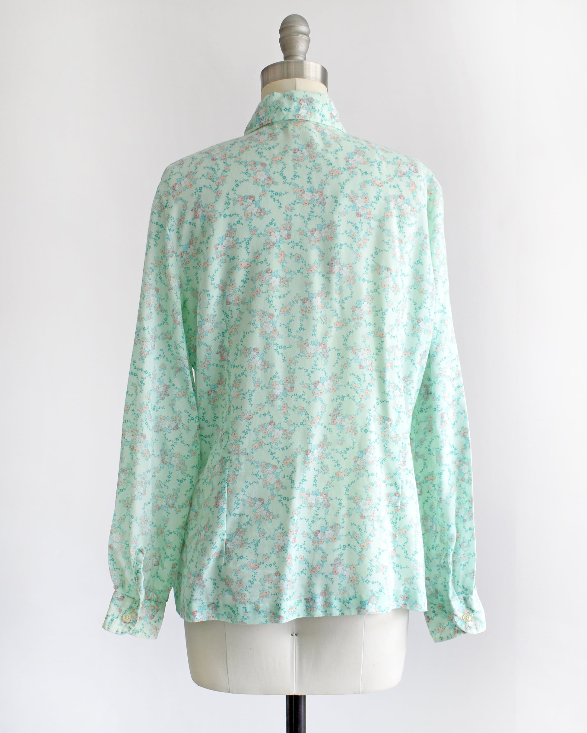 back view of a vintage 1970s long-sleeve button-up blouse with a light mint green and pink floral pattern, displayed on a cream dress form mannequin against a white background.