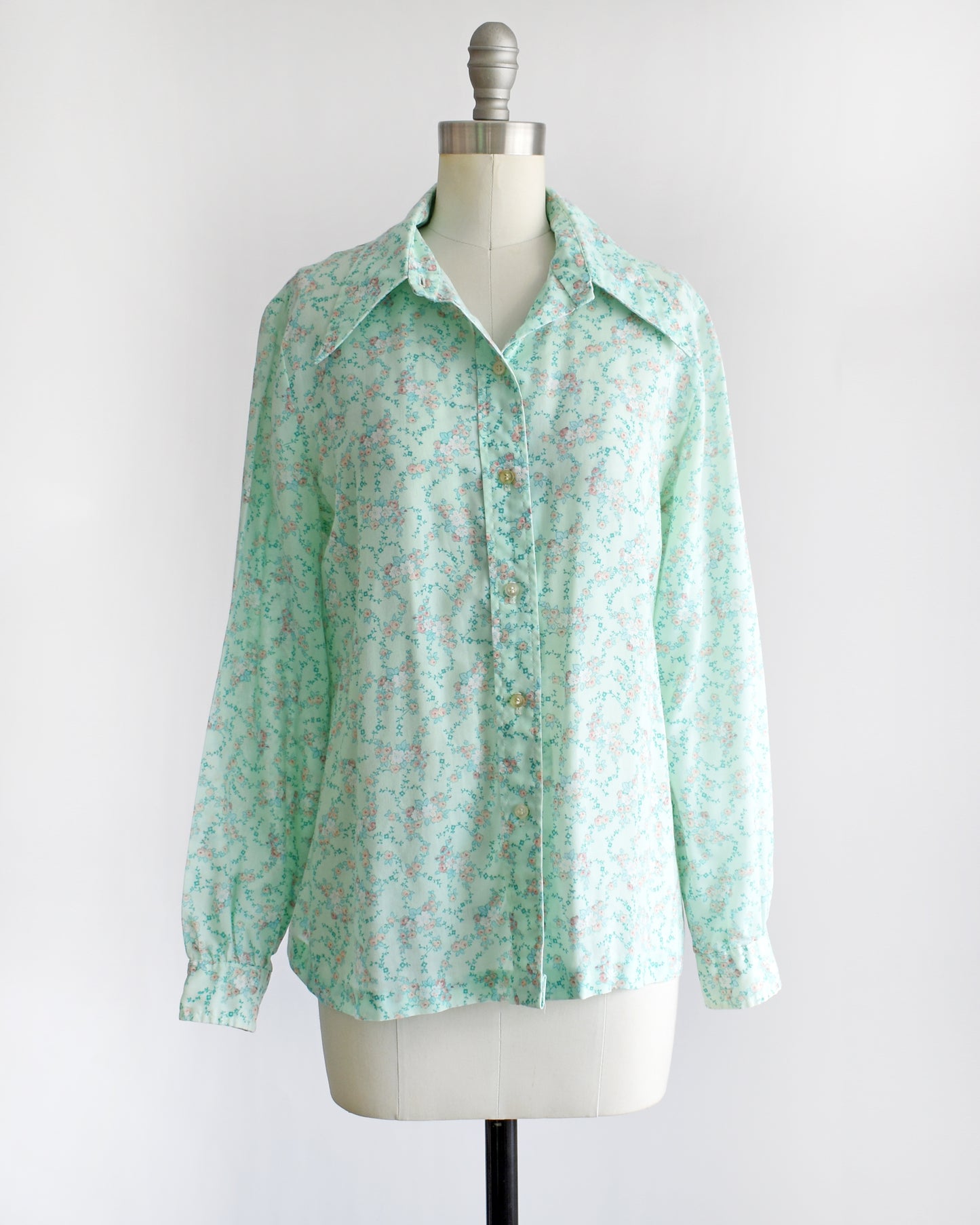 a vintage 1970s long-sleeve button-up blouse with a light mint green and pink floral pattern, displayed on a cream dress form mannequin against a white background.