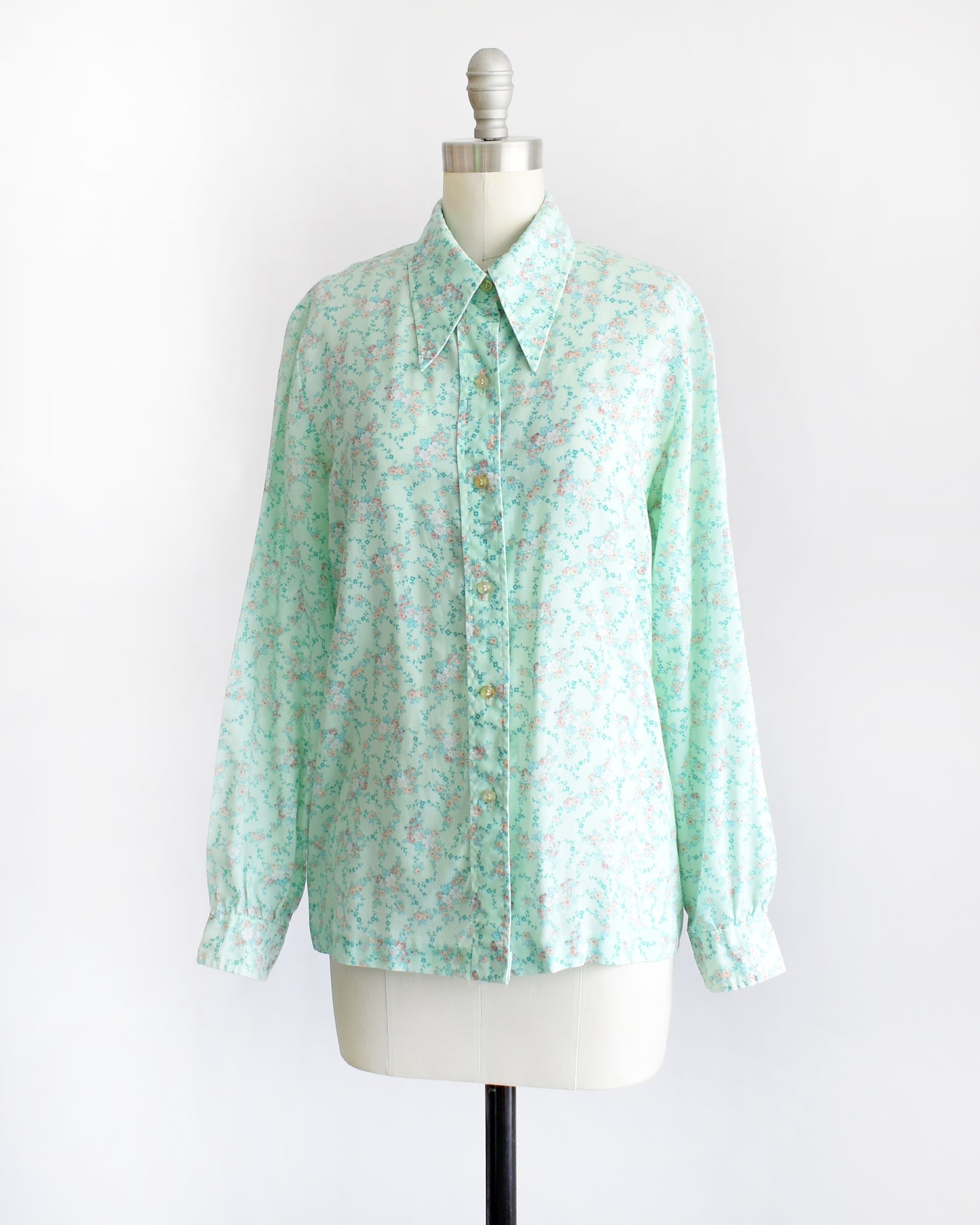 side front view of a vintage 1970s long-sleeve button-up blouse with a light mint green and pink floral pattern, displayed on a cream dress form mannequin against a white background.