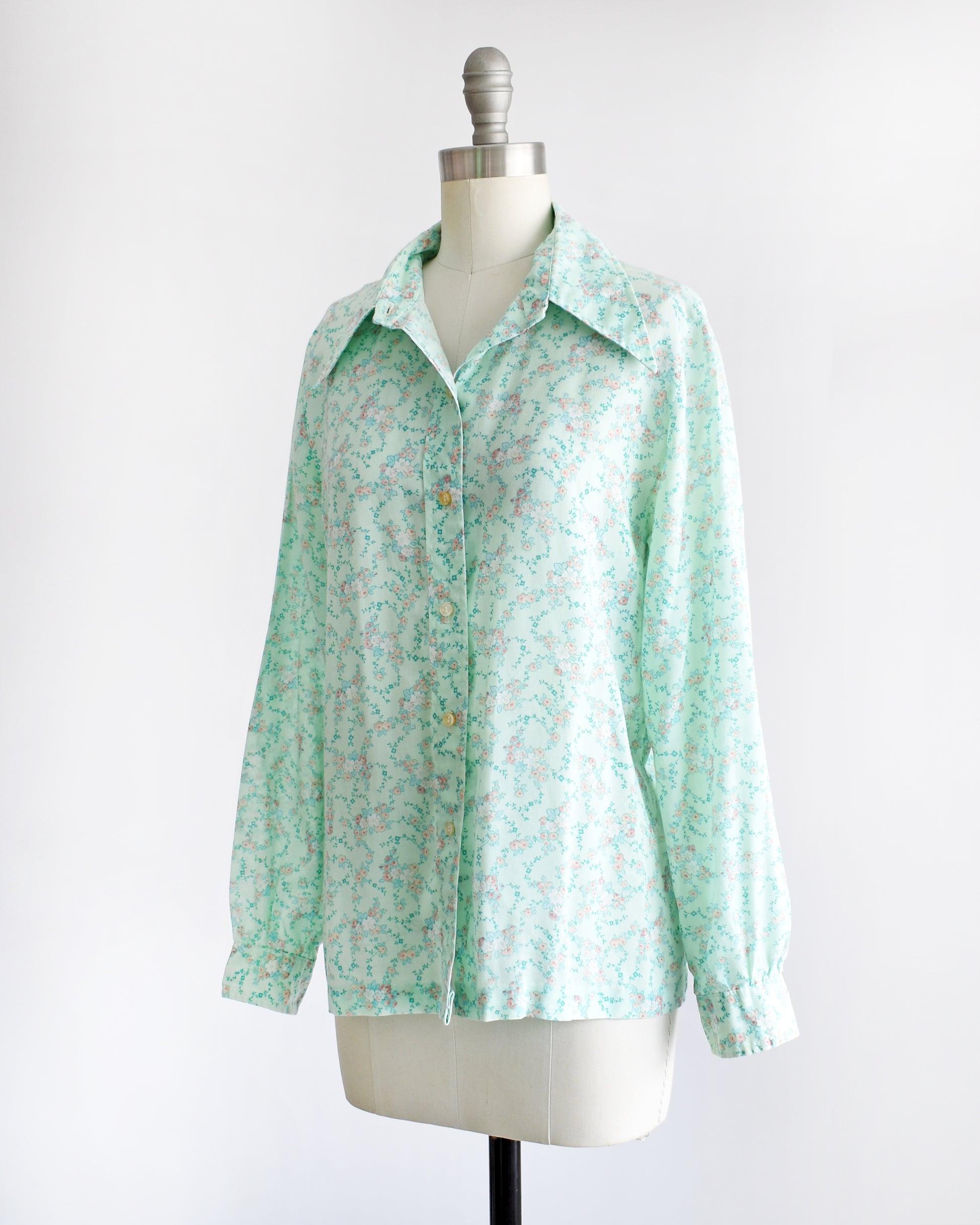 side front view of a vintage 1970s long-sleeve button-up blouse with a light mint green and pink floral pattern, displayed on a cream dress form mannequin against a white background.