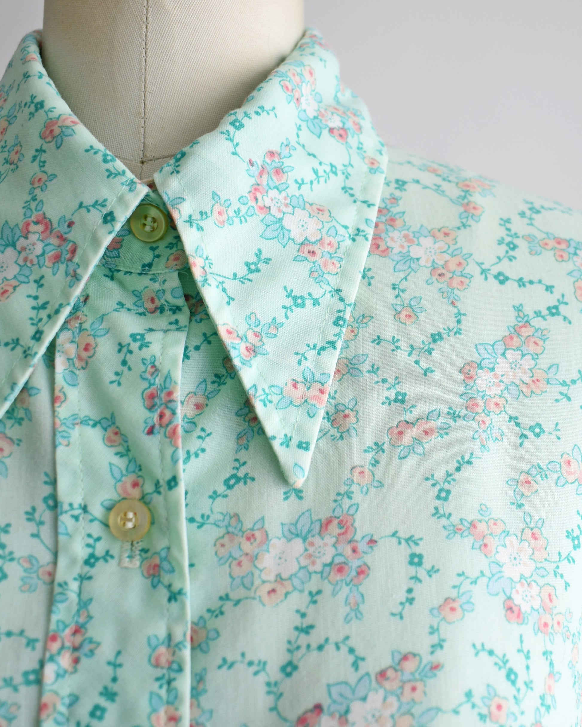A close-up of a vintage collared blouse on a mannequin, featuring a light mint green base with a delicate pink and white floral print and green buttons.