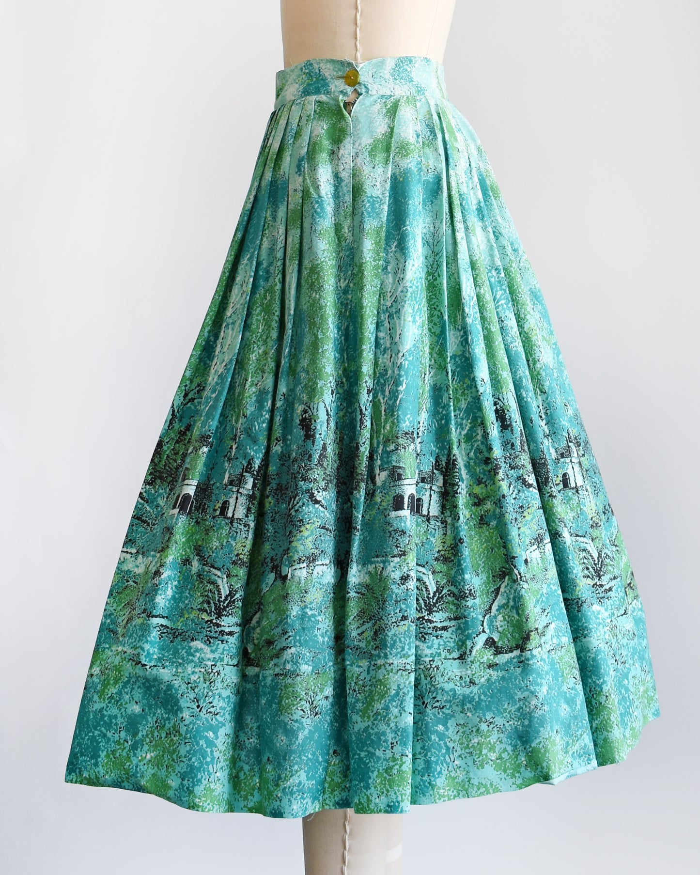 side view of  vintage 1950s blue and green novelty print skirt that has a scenic forest and chateau border print scene.