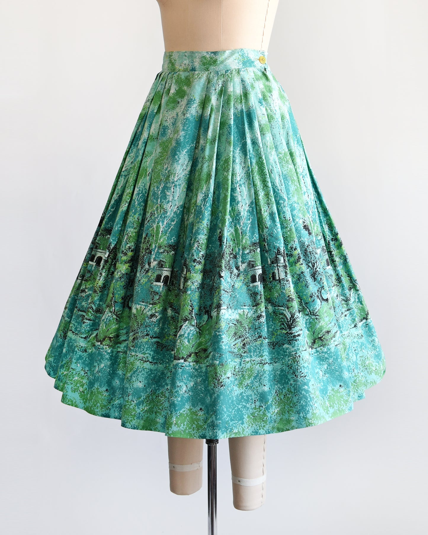 side front view of a vintage 1950s blue and green novelty print skirt that has a scenic forest and chateau border print scene.