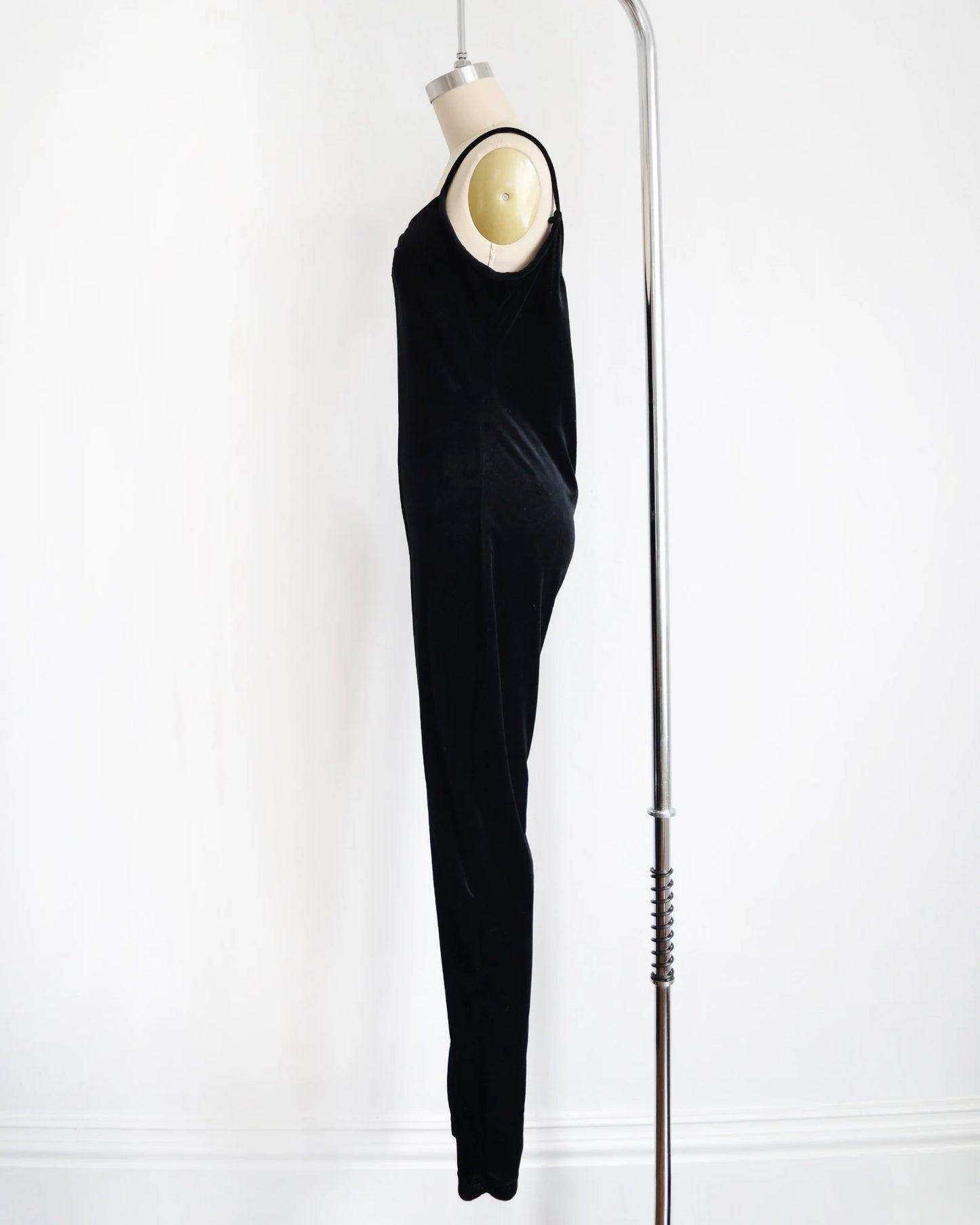 side view of ablack velvet jumpsuit displayed on a mannequin against a white wall, with a silver bar stand visible on the right side.