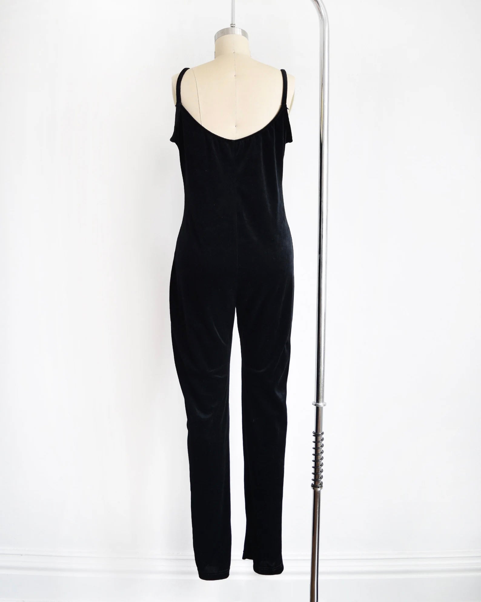 back view of a black velvet jumpsuit displayed on a mannequin against a white wall, with a silver bar stand visible on the right side.