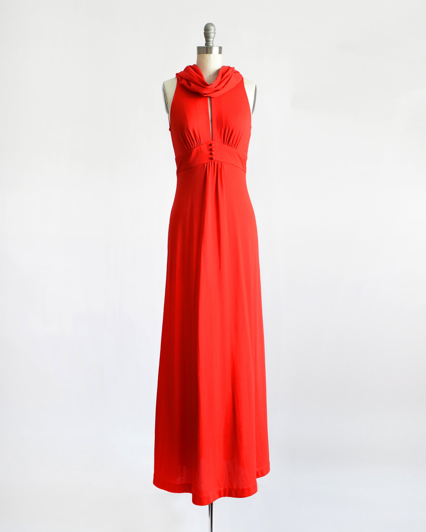 A bright vintage 1970s red sleeveless maxi dress with a draped cowl neckline and a front button detail, displayed on a dress form mannequin against a plain white background.
