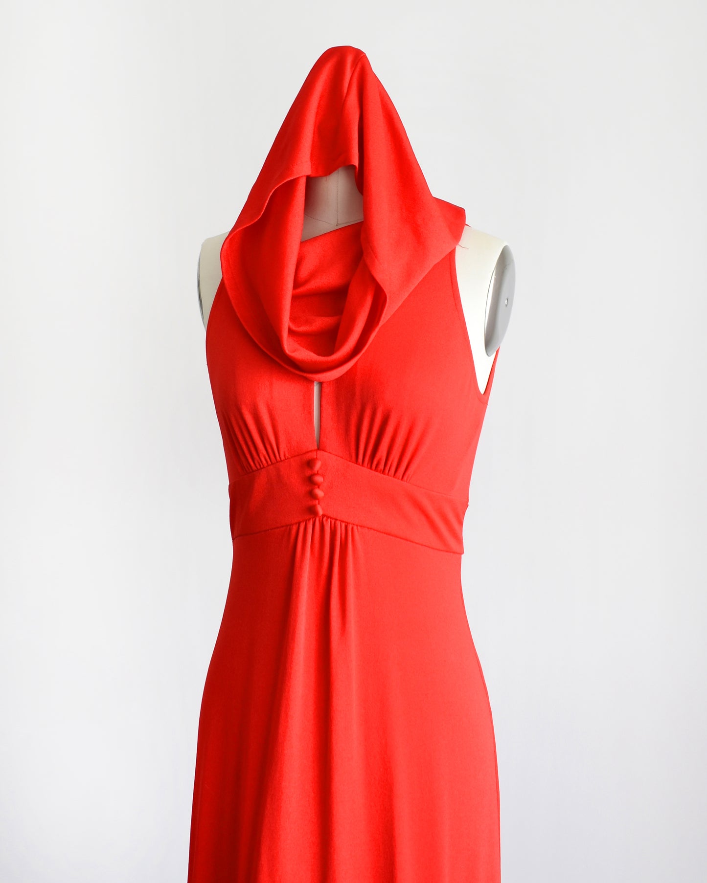 A bright red sleeveless halter-neck dress with a draped cowl hooded neckline and gathered waist, displayed on a white mannequin against a plain white background.