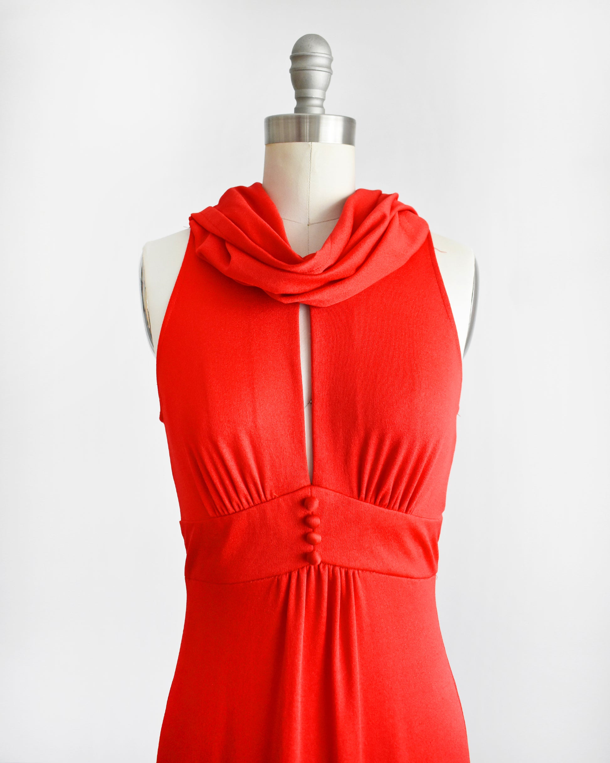 A bright red sleeveless dress displayed on a cream dress form mannequin against a light gray studio background. The dress features a draped cowl neckline, a front keyhole cutout with a tie, gathered detailing at the waist with three decorative buttons, and a flowing skirt.